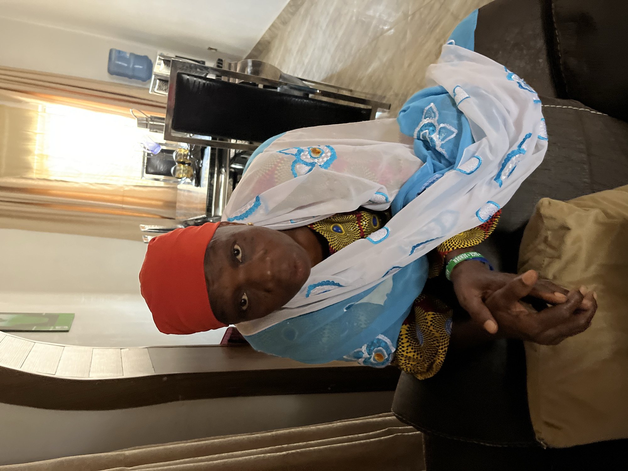An indoor portrait photograph, taken in what appears to be a home in Jos, Nigeria, features an older woman seated on a dark brown sofa, looking directly at the camera. She wears a red headscarf and a brightly patterned top or dress in yellow, black, and blue, featuring circular motifs. A light blue and white sheer scarf, adorned with intricate white and blue floral embroidery, is draped around her. On her left wrist, she has a green wristband with the partially visible text "ACK SENDER."
The setting is a well-lit room, with bright natural light entering from a window covered by horizontal blinds in the background, suggesting it is daytime. Behind her, there is modern furniture with metallic frames, possibly a dining table or counter, on which a blue water dispenser and other items are visible. Her hands are clasped gently in front of her. The overall ambiance is that of a comfortable, contemporary living space.