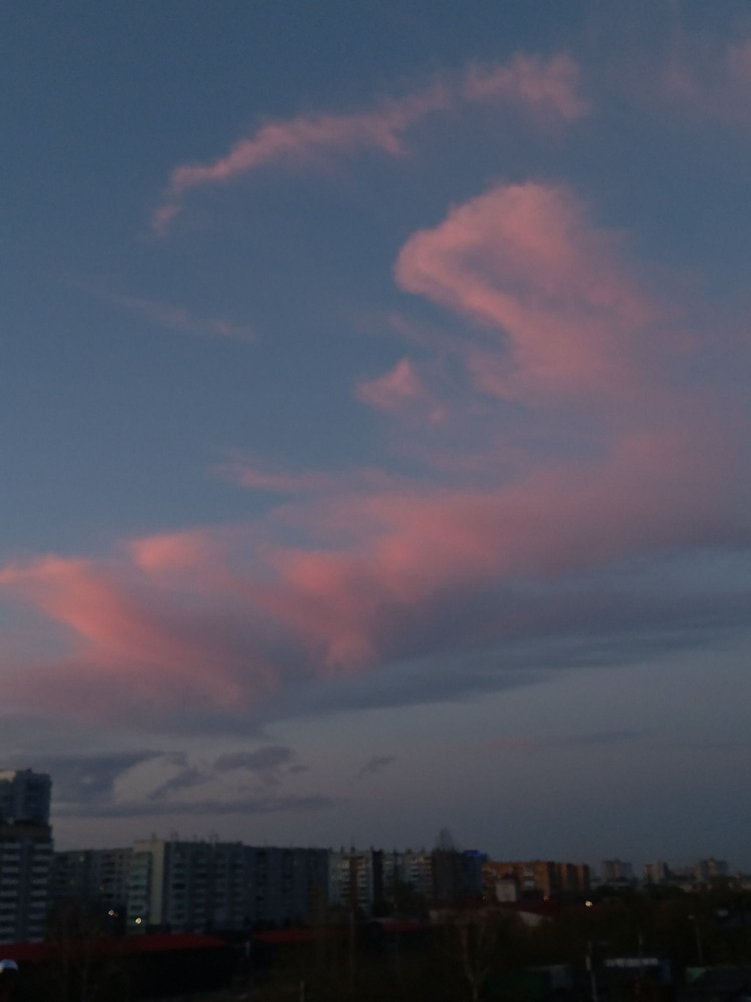 The image shows a cityscape at dusk, likely in Krasnoyarsk, Russia, although specific location cannot be confirmed without additional information. The sky occupies most of the frame, featuring pink and dark gray clouds against a blue background. Below, a line of buildings dominates the horizon, with multiple structures and some visible trees. The image is slightly blurry.