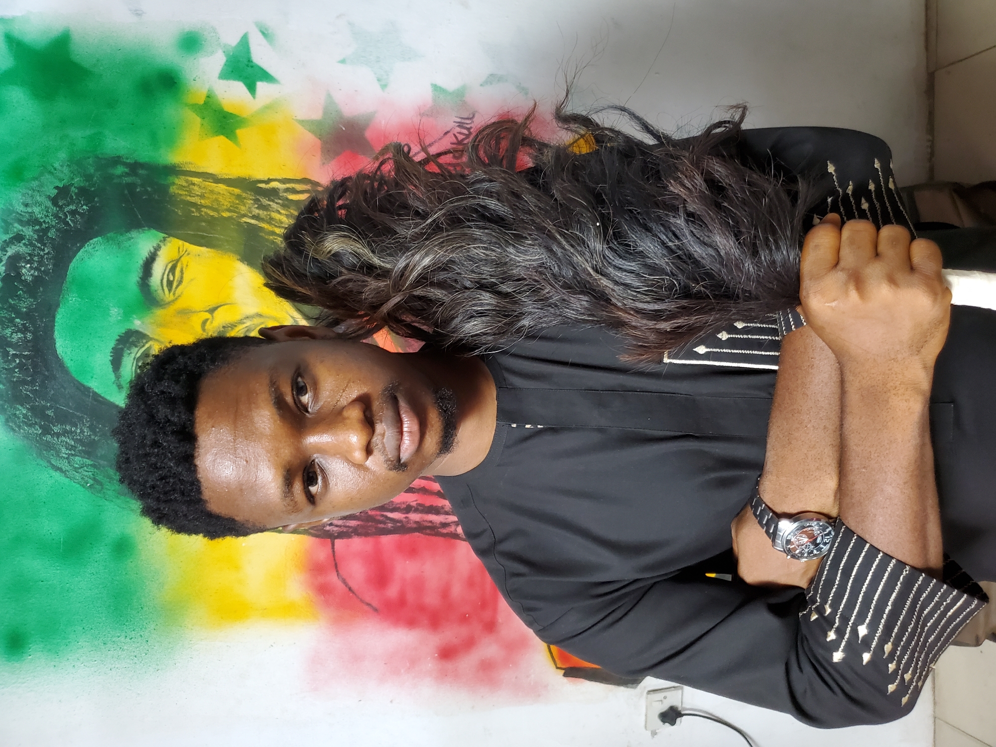 A man with dark skin and short, dark hair stands in front of a wall with a painted mural in a city. The mural is vertically oriented and features a portrait of a person with skin tones in green, yellow, and red, with star-shaped stencils in the background. The mural may have been done with spray paint. The man wears a long-sleeved black shirt with geometric gold embellishments on the cuffs and on the front. He has a watch on his left wrist and his arms are crossed. The area where the picture was taken is likely in Mararaba, Nigeria.