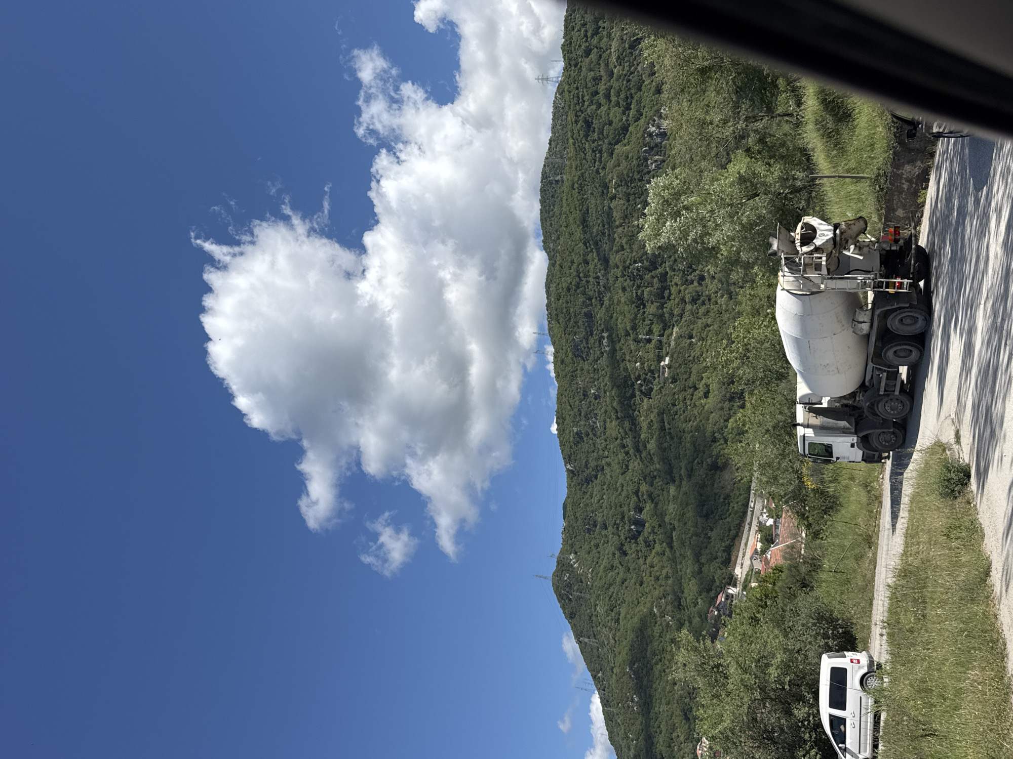 The scene is located in Podlastva (Poljice), Montenegro. The image depicts a view of a landscape featuring a blue sky with a large cloud formation, a green mountainous area, and a road with vehicles. A concrete mixer truck is parked on the side of the road. Further down the road and slightly obscured by trees is a white van. Houses can be seen in the mid-ground, nestled near the base of the mountain. Electrical pylons are on the mountain. The image appears to have been taken from inside a vehicle due to the dark frame at the top of the image.