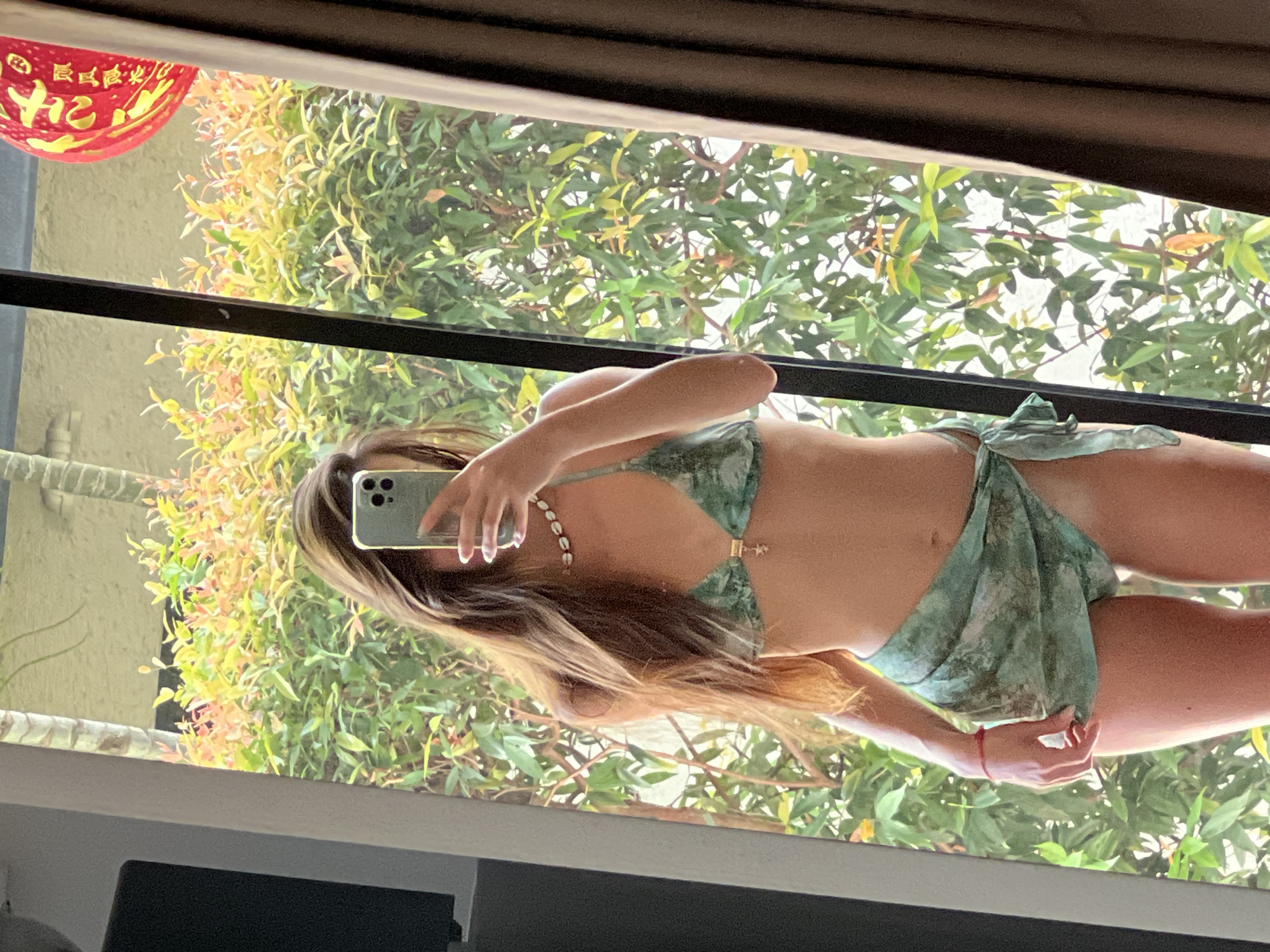 A young woman with long brown hair is reflected in a mirror, taking a selfie with her phone. She is wearing a green patterned bikini and a matching sarong tied around her waist. She is also wearing a seashell necklace. Behind her, the reflection shows lush green foliage and a red lantern hanging on the right side. The setting appears to be outdoors, possibly in a tropical or warm climate, with the reflection indicating a natural backdrop.