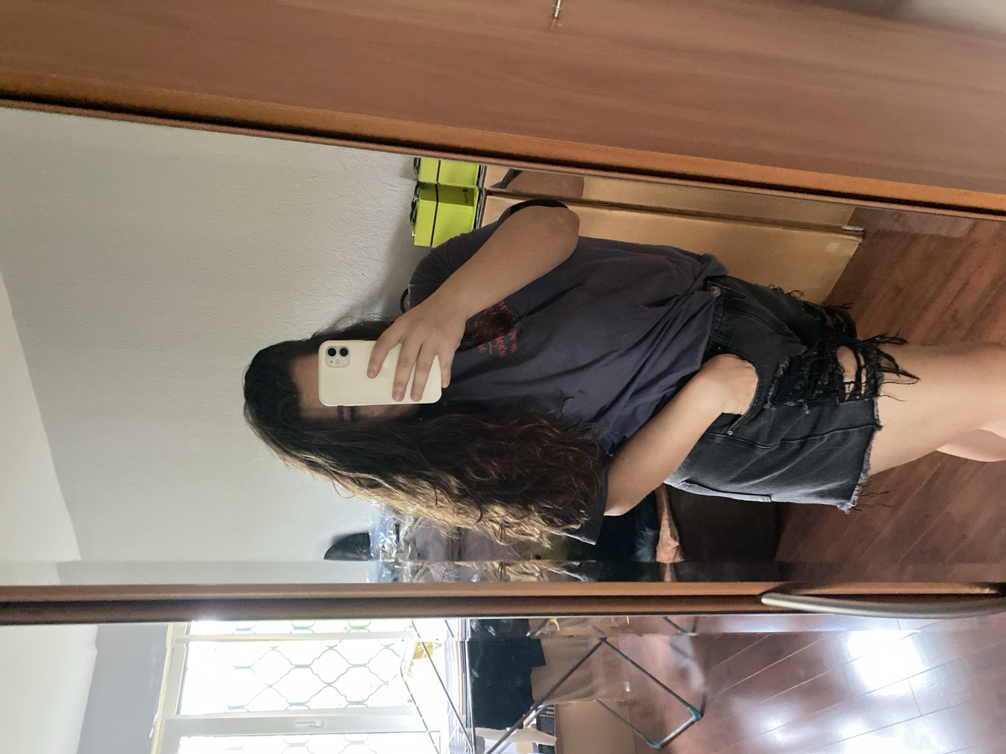 A person is taking a mirror selfie in what appears to be a bedroom. The person has long, wavy, dark brown hair and is wearing a dark grey t-shirt and distressed black denim shorts. Their left hand is in their pocket. The mirror reflects a window with a patterned screen, a clothes drying rack with laundry, cardboard boxes, and a wooden floor. In the reflection of the room, there are two bright green boxes on a surface, and a wooden closet door. The image does not provide any specific information to locate it within the city of Perm, Russia.