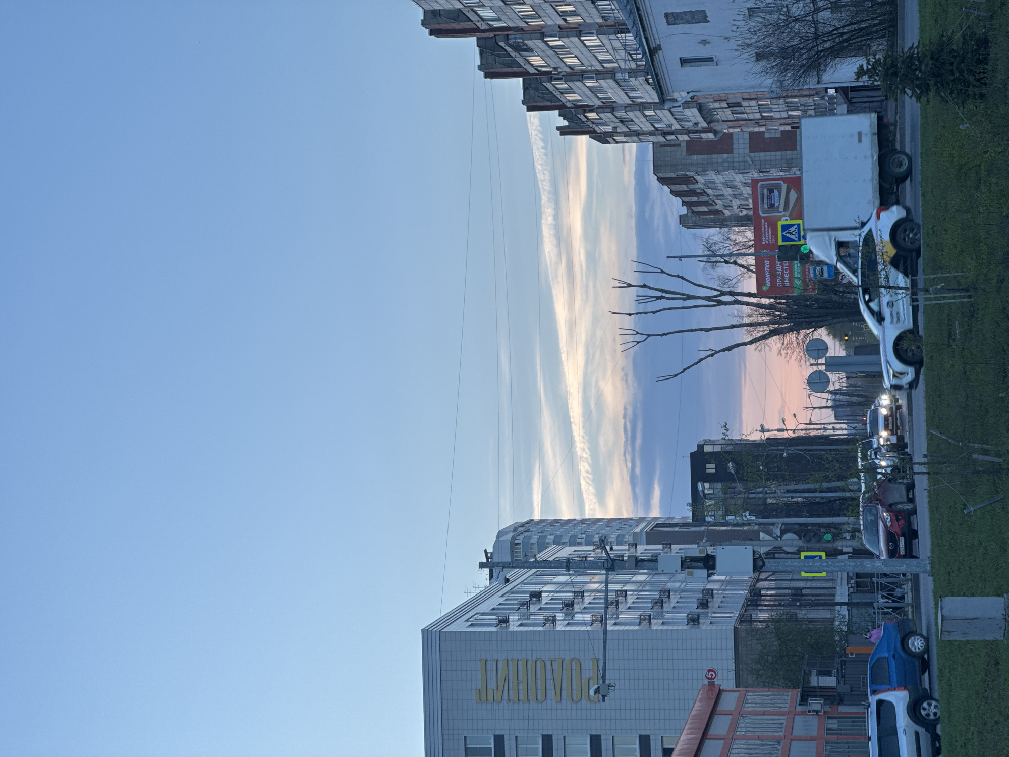 The image captures a street scene in Perm, Russia, showcasing a clear, blue sky with linear clouds. Buildings line both sides of the street, a mix of residential and commercial structures. A sign in Cyrillic (РОАОИЛНТ) is visible on one building, with other advertisements along the roadway. The foreground features parked cars, traffic lights, and street signs. Cars are also visible further down the street, suggesting traffic. Bare tree branches are silhouetted against the sky, indicating either early spring or late autumn. The overall scene conveys a calm urban environment with typical street furniture and infrastructure.