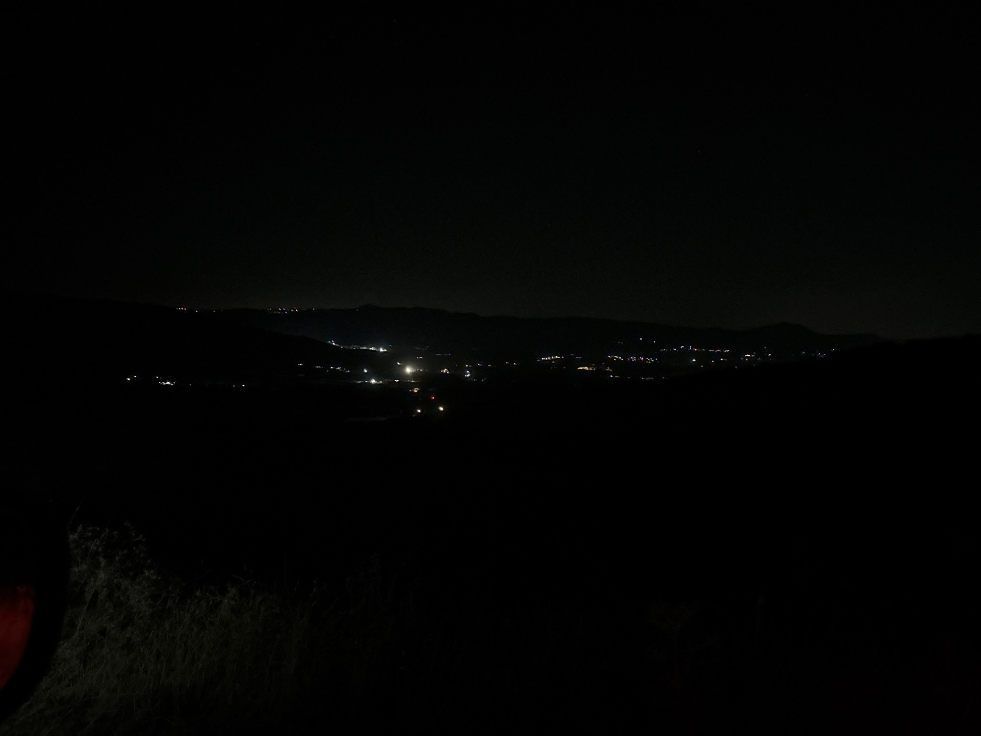 The image depicts a nighttime aerial view of a landscape with scattered lights indicating a settlement. The foreground is dark, with some textured vegetation visible in the lower left corner. In the middle ground and background, a series of lights are spread across a dark, hilly terrain, suggesting the presence of buildings and infrastructure within the Armenochori Community in Cyprus. The sky is dark and appears clear, with no visible celestial bodies or significant atmospheric phenomena. The overall impression is one of a tranquil, sparsely populated area at night.