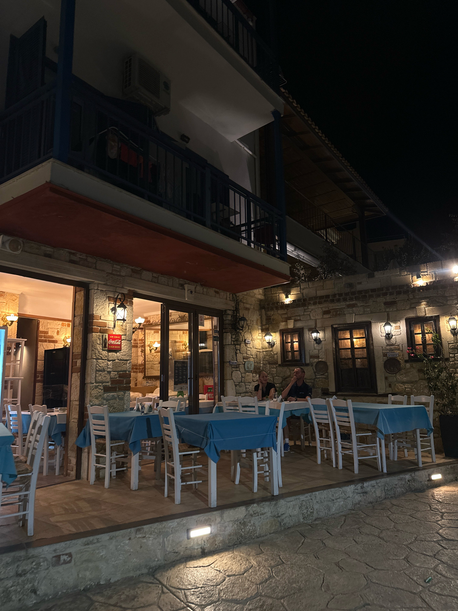 The image shows a nighttime exterior view of a restaurant located in Kassandra Municipal Unit, Greece. The restaurant has a stone facade with large glass doors and windows, and tables are set with blue tablecloths and white chairs both inside and outside. An air conditioning unit is visible on the upper level balcony, which has a blue railing. Two patrons are seated at a table on the patio, a man and a woman. The lighting is artificial, casting a warm glow on the scene.