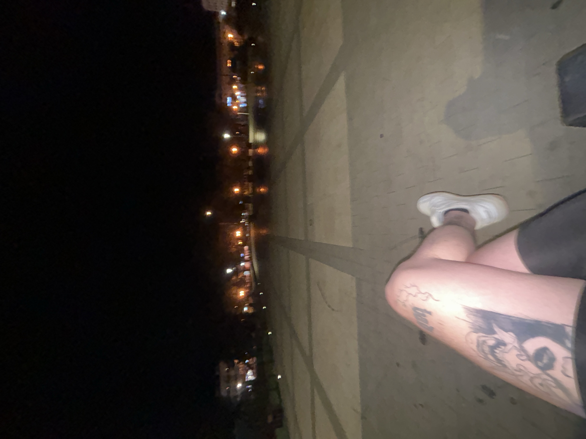 The image depicts a low-angle, night-time perspective from a seated position, focusing on the photographer's tattooed left leg and white athletic shoe. The leg exhibits a prominent tattoo of a creature with sharp teeth, possibly a dragon or monster, extending down the thigh. The background reveals a city square or promenade in Siedlce, Poland, characterized by patterned paving stones, streetlights casting warm glows, and illuminated buildings in the distance. The ground appears slightly wet, reflecting the lights, suggesting recent rain or damp conditions. The overall scene is dark, with artificial lighting creating a moody atmosphere, and the focus is on the personal element of the tattoo and the broader urban environment.