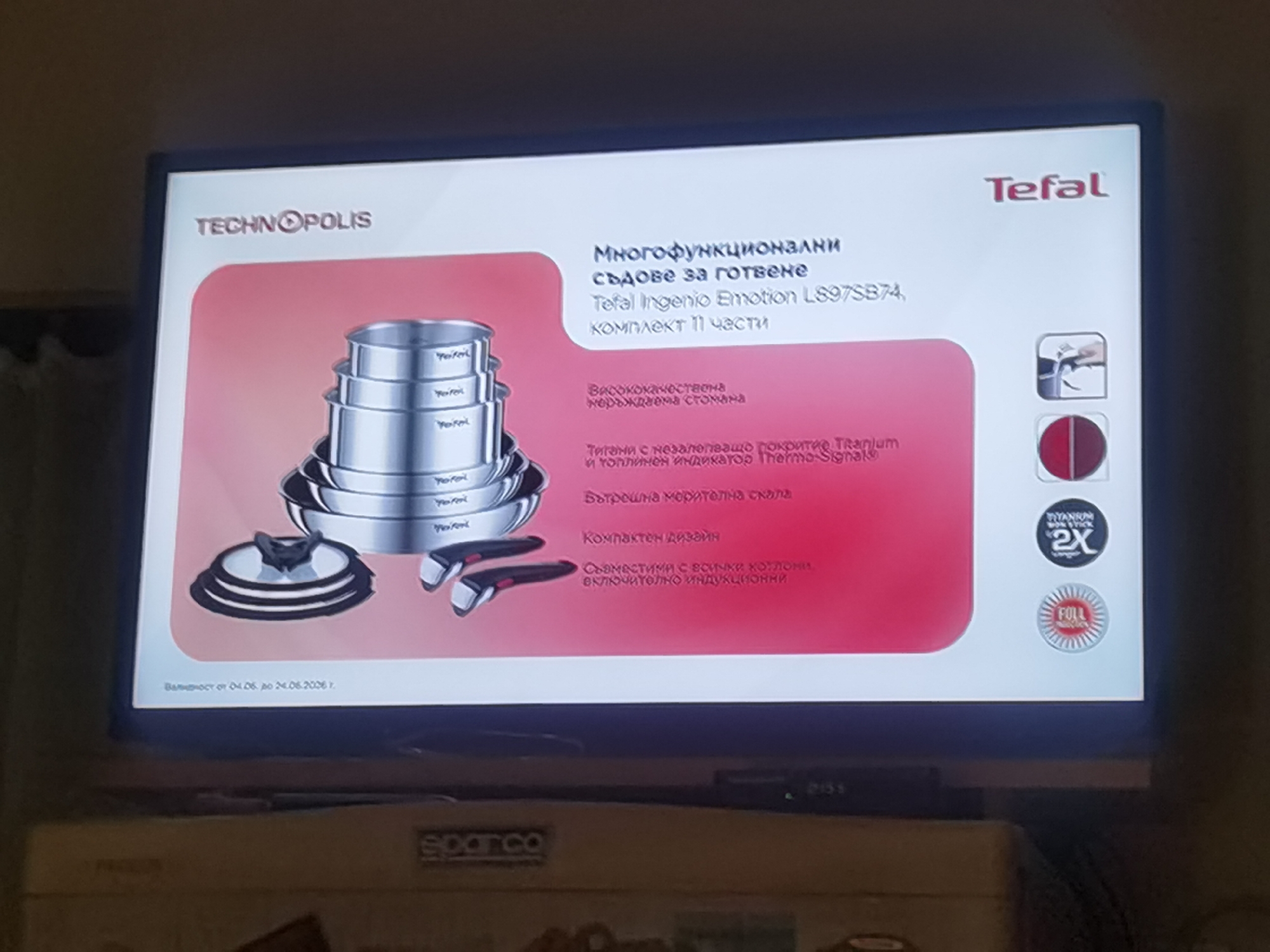 The scene depicts a television screen displaying a commercial advertisement, located in what is identified as the city of Lomets, Bulgaria. The screen occupies the majority of the frame, showing a white background with a large pink/red section.
At the top left of the screen, the "TECHNOPOLIS" logo is visible. At the top right, the "Tefal" logo is displayed. The advertisement showcases a Tefal Ingenio Emotion L897SB74 11-piece cookware set, consisting of several stainless steel pots and pans of varying sizes, stacked concentrically, along with a transparent lid with a black handle and two detachable black handles.
Bulgarian text on the screen details the product's features: "Многофункционални съдове за готвене" (Multifunctional cooking vessels), "Висококачествена неръждаема стомана" (High-quality stainless steel), "Тигани с незалепващо покритие Titanium и топлинен индикатор Thermo-Signal" (Pans with Titanium non-stick coating and Thermo-Signal heat indicator), "Вътрешна мерителна скала" (Internal measuring scale), "Компактен дизайн" (Compact design), and "Съвместими с всички котлони, включително индукционни" (Compatible with all hobs, including induction). An expiration date for an offer is visible at the bottom left, reading "Валидност от 04.06. до 24.06.2021" (Validity from 04.06. to 24.06.2021).
Accompanying icons on the right side of the screen illustrate features: a hand detaching/attaching a handle, a red circular "Thermo-Signal" indicator, a "TITANIUM NON-STICK 2X" logo, and a "FULL INDUCTION" logo.
Below the television, a piece of furniture is visible, bearing the "SPARCO" brand logo. The immediate environment is an indoor setting. No individuals are present in the image.