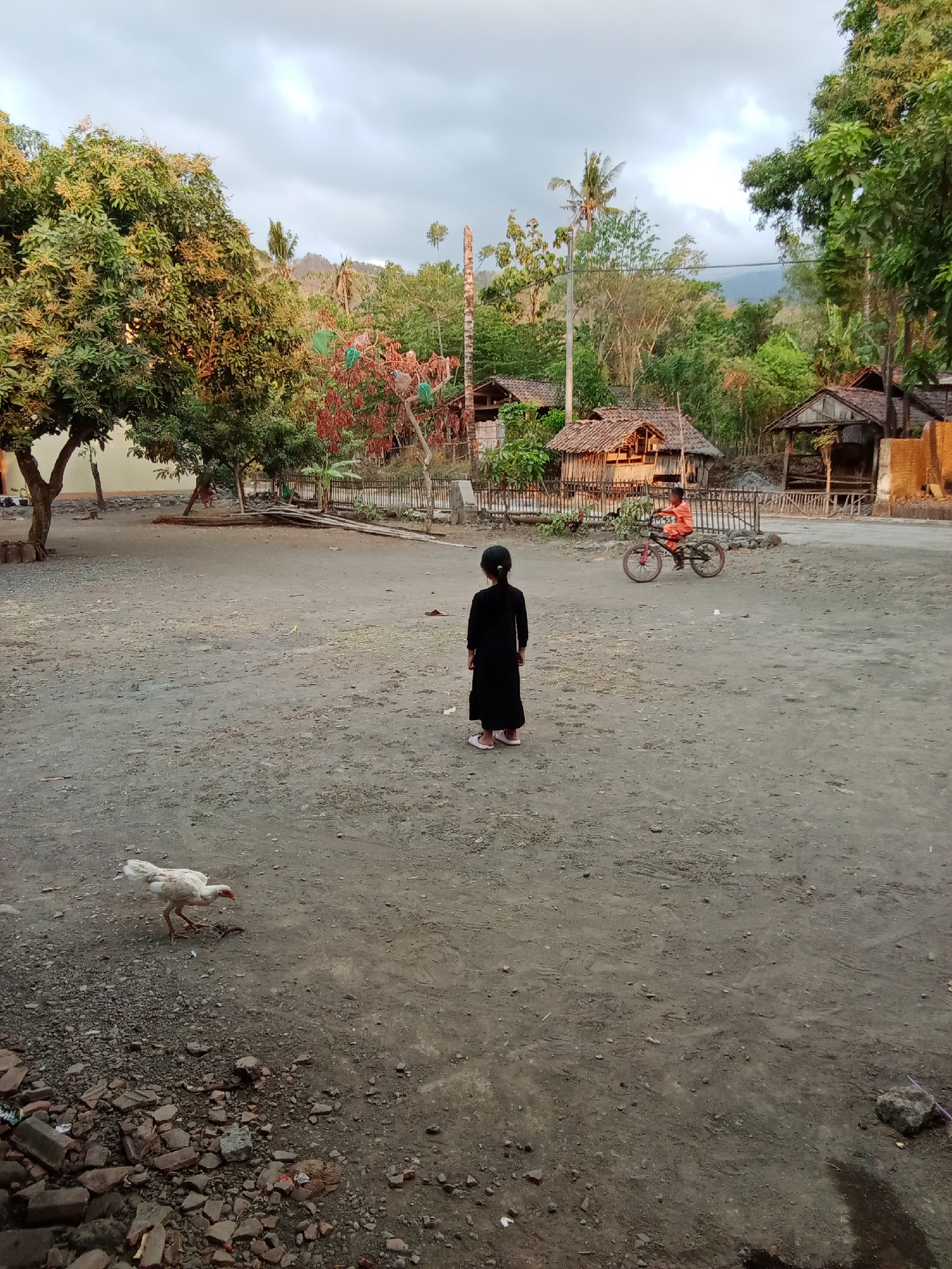 A wide-angle, low-light, full shot captures a dirt yard in Selowogo, Indonesia. A young girl in a black dress and pink sandals stands with her back to the camera, facing a dirt path. To her left, a white chicken pecks at the ground near scattered rocks and debris. In the background, on the right side of the yard, a child wearing an orange shirt rides a bicycle. Behind the children, several simple wooden houses with tiled roofs are visible, nestled among lush green trees and foliage, with a gently sloping hill in the distance under a cloudy sky.