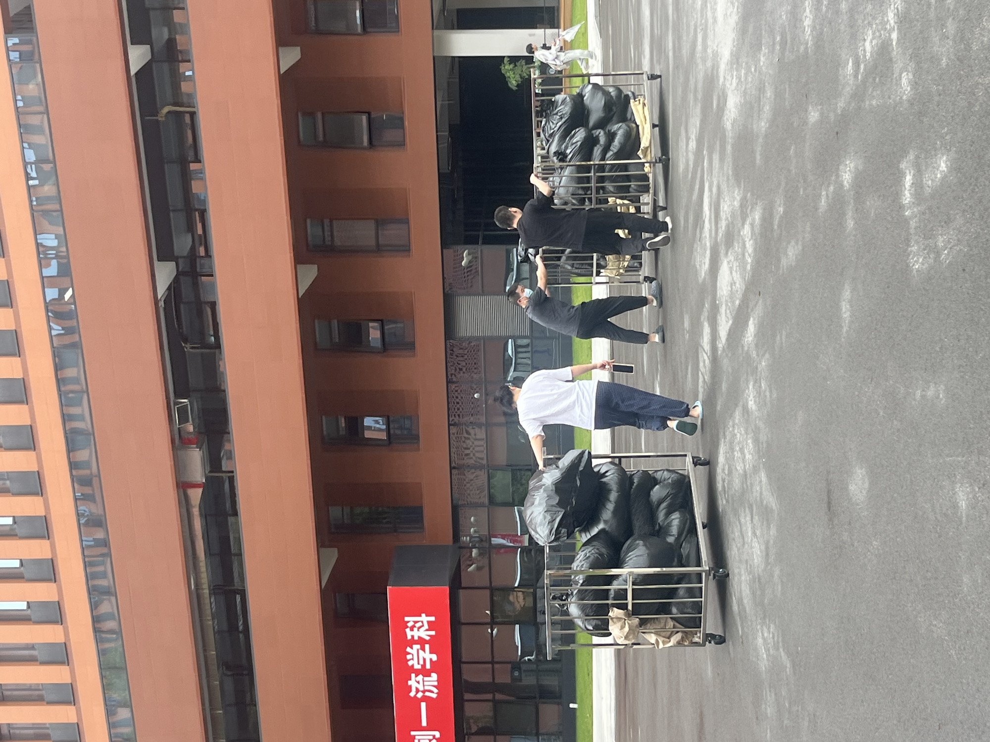 This is a medium shot of three men and one person in the background near a modern building, likely in Yuzhong District, China. In the foreground, two men are pushing a large metal trolley laden with numerous black trash bags. The man on the left wears a dark t-shirt, dark pants, and a light-colored mask, while the man on the right wears a black t-shirt and black pants. To the left of this trolley, another trolley is similarly loaded with black trash bags, and a man in a white t-shirt and patterned pants walks away from it, looking back. In the background, a person wearing white is visible to the far right, holding a white object, possibly a broom or mop. The building behind them is modern, with a reddish-brown facade and multiple rows of dark windows. A large red sign with white Chinese characters is mounted on the building to the left. The ground appears to be asphalt or concrete.