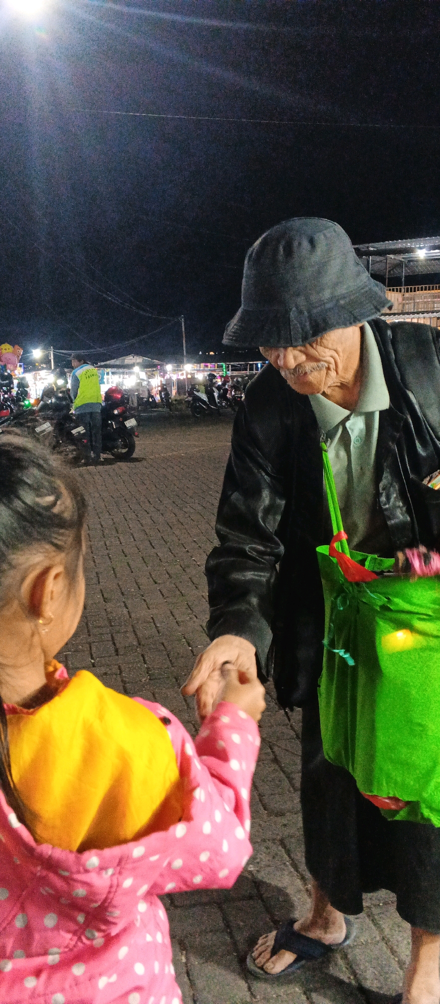 This is a vertical, medium shot photograph taken at night in what appears to be an outdoor market or event in Karangploso, Indonesia. The lighting suggests it is nighttime, with artificial lights illuminating the scene.
The primary subjects are an elderly man and a young girl. The man is wearing a dark hat, a light-colored collared shirt, and a dark jacket. He has a mustache and appears to be looking down and slightly to his right, extending his right hand. He is also carrying a large, bright green bag with neon green straps. The young girl, seen from the back, is wearing a pink hooded jacket with white polka dots, and a bright yellow inner layer is visible. She is facing the man and reaching out her left hand to his. Their hands are almost touching, suggesting an interaction like a handshake or the passing of an object.
In the background, there are more people, motorcycles, and stalls or temporary structures, consistent with a market or festival setting. Some of the stalls are lit, and balloons are visible on the left side of the frame. A person in a yellow vest is standing near some motorcycles. The ground is paved with interlocking bricks.
The overall mood seems warm and possibly sentimental, given the interaction between the elder and the child. The night setting, the lighting, and the crowded market environment contribute to a lively, perhaps festive atmosphere. No visible text is discernible in the image.