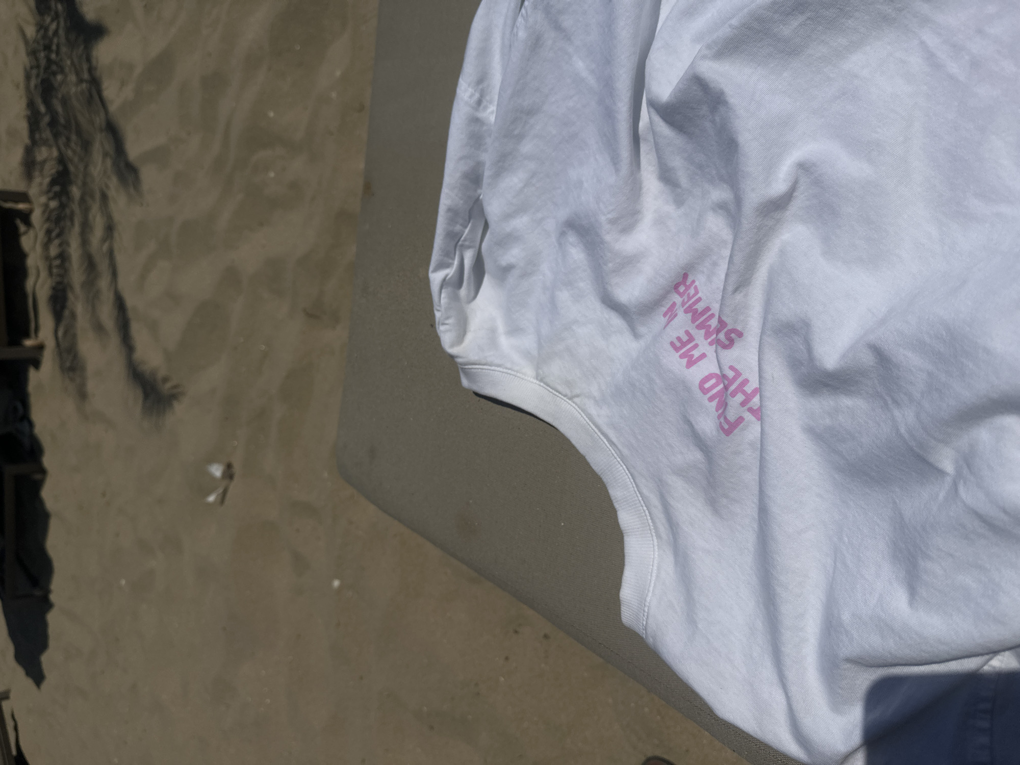 A white t-shirt with pink lettering that reads "FIND ME IN SUMMER" is draped on a tan-colored surface. The t-shirt is lying on a beach, as evidenced by the sand in the background. Palm tree shadows are visible on the sand, suggesting a warm, sunny day. The scene is likely in Nesebar, Bulgaria, given the context.