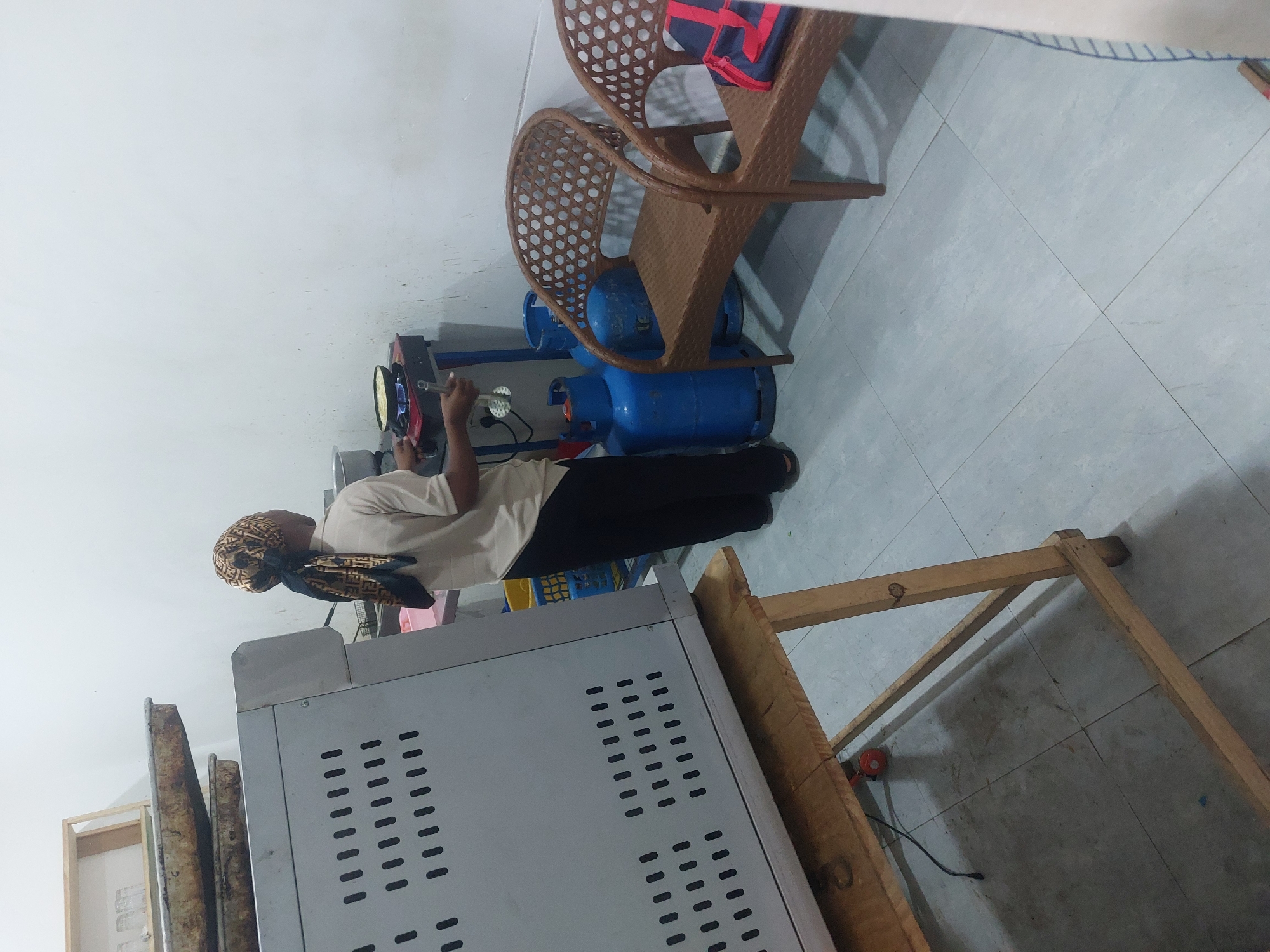A person wearing a headscarf stands in what appears to be a kitchen or food preparation area. They are near a large, stainless steel oven. In the background, there are two blue propane tanks, a cooking appliance with pans, and brown wicker chairs. The floor is tiled and the walls are white. Based on the presence of propane tanks, it is likely an area where food preparation is conducted. Without additional information, it is not possible to confirm the specific location within Dar es-Salaam, Tanzania.