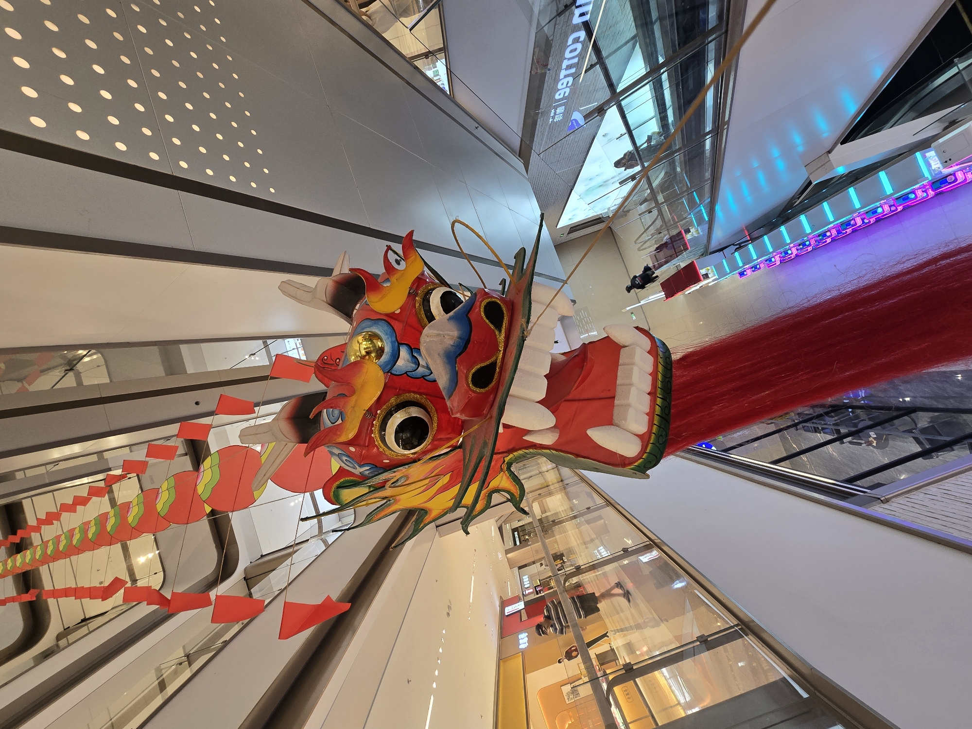 A large, red dragon head decoration, with yellow and blue accents and wide open jaws revealing white teeth, hangs from the ceiling of a multi-level shopping mall in Xiaoshan District, China. Red and circular red and green ornaments are strung above and behind the dragon. The mall features escalators with bright blue and purple lights, glass railings, and shops visible on the lower levels. Two people are seen walking on the escalators and another person is walking on the second level. The ceiling is made of white panels with a pattern of circular cutouts through which light emanates.