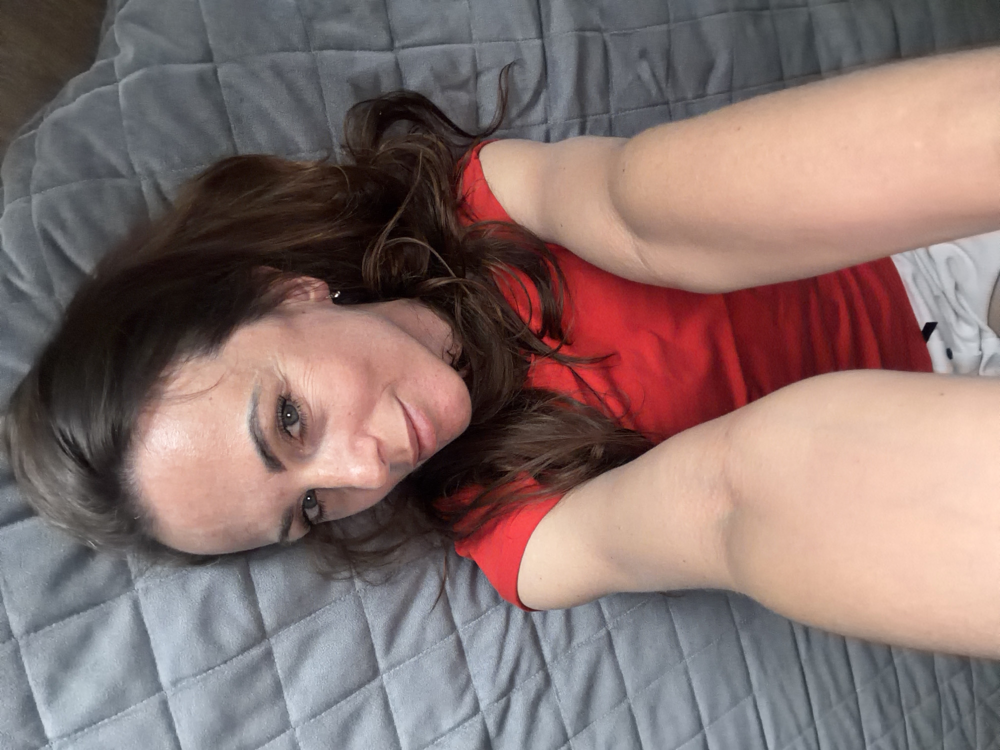 A woman with shoulder-length brown hair is lying on a grey quilted blanket. She is wearing a red t-shirt and white shorts. The location is unspecified and not discernable from the image. Birobidzhan, Russia is not evident in the image.