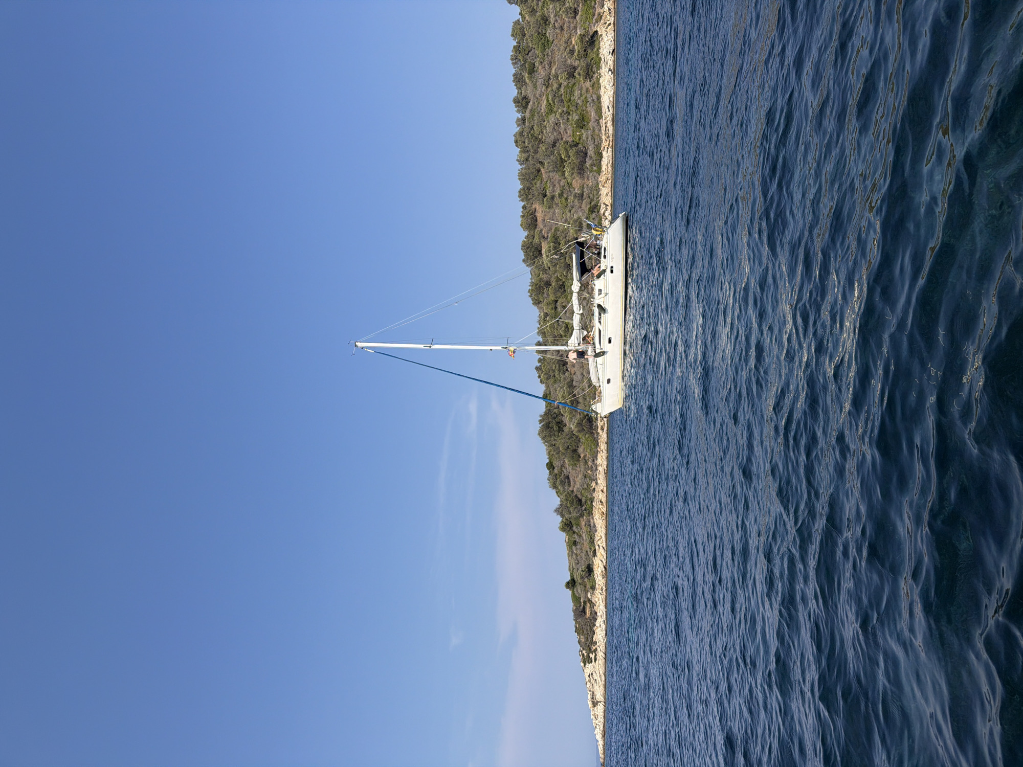 An aerial view of a sailboat on the Aegean Sea, off the coast of an island in Greece. The sailboat is a white catamaran with a blue sail. It is anchored in the water, and there are people on deck. The island is covered in olive trees and rocks. The sky is clear and blue. The water is deep blue and calm, with small ripples on the surface. The scene is peaceful and serene, evoking a sense of relaxation and tranquility.