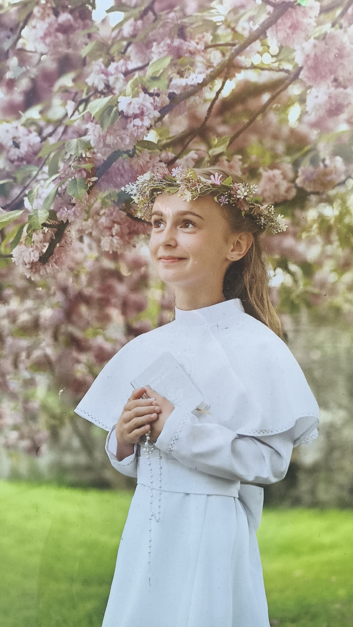 A young girl stands in a garden or park, likely in Nowy Tomyśl, Poland, based on local customs regarding religious events. She is dressed in a white ceremonial robe and cape, indicating a First Communion or similar religious occasion. She wears a floral crown, holding a small prayer book with Polish text and a rosary. Pink blossoms are in the tree canopy above her. Green grass is in the background.