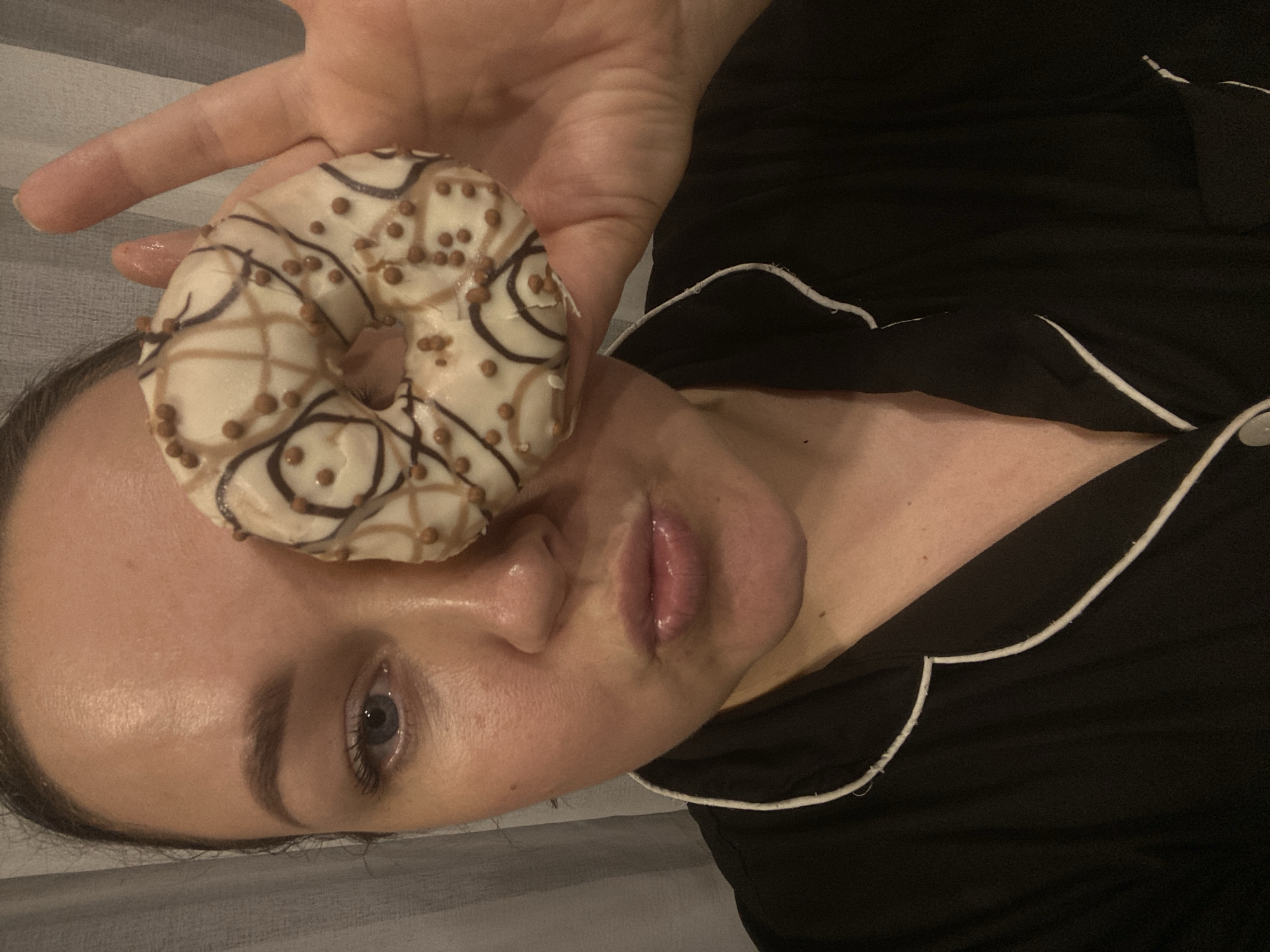 The image presents a close-up, chest-up portrait of an adult female in an indoor setting. The subject's left eye, nose, mouth, and portions of her forehead and chin are visible, with her lips pursed. She is wearing a black garment with white piping, consistent with a pajama top. Her right hand holds a single donut, positioned directly over her right eye, completely obscuring it. The donut features a white glaze, dark brown and light brown drizzles, and is topped with numerous small, spherical, light brown sprinkles. The background consists of a light-colored, vertically textured fabric, likely a curtain or drapery. No specific visual information within the image identifies the location as Sochi, Russia.