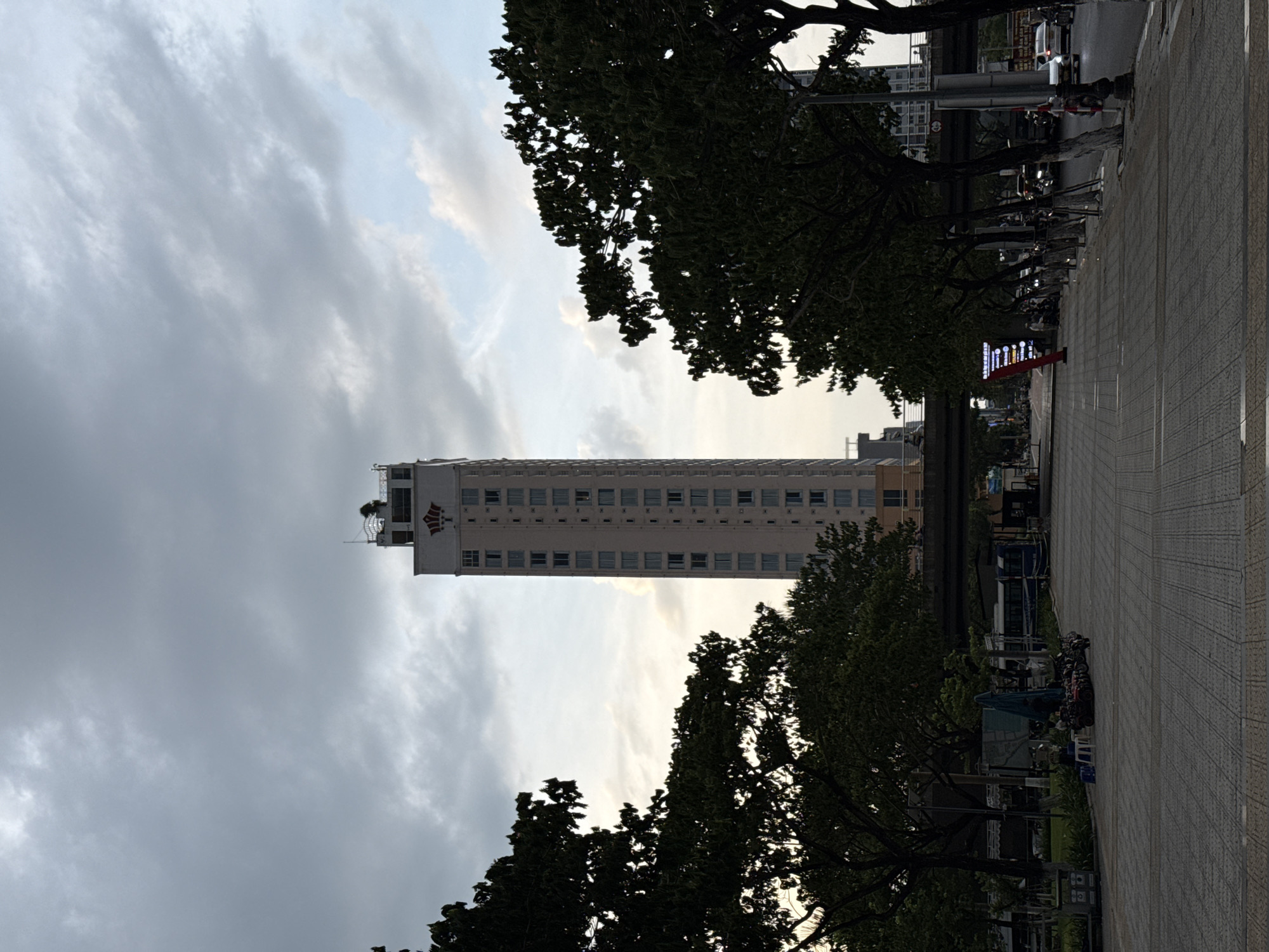 This is a medium shot taken from a ground-level perspective in Đà Nẵng, Vietnam, looking down a paved walkway toward a tall, slender, light-colored building. The building has a distinctive logo near its top which appears to be a stylized crown. The sky above is overcast with dramatic clouds, hinting at an impending storm or recent rain. On either side of the walkway, large, dark green trees with dense foliage create a canopy, framing the view of the building.
In the foreground, the walkway is paved with light-colored square tiles arranged in a diamond pattern. To the left of the walkway, there is a grassy area with some sparse vegetation and a few parked motorcycles. A blue tarp is propped up with a stand. A few people are visible sitting on small stools near the motorcycles. Further down the walkway, a large digital billboard with white text on a dark background is positioned.
Beyond the walkway and trees, a bridge or overpass spans across the scene, with traffic visible underneath. On the right side of the image, a road runs parallel to the walkway, with several cars and motorcycles in motion. Streetlights and traffic signals are also present. The overall impression is a city scene during a cloudy day, possibly late afternoon or early evening, with a mix of urban infrastructure and natural elements.