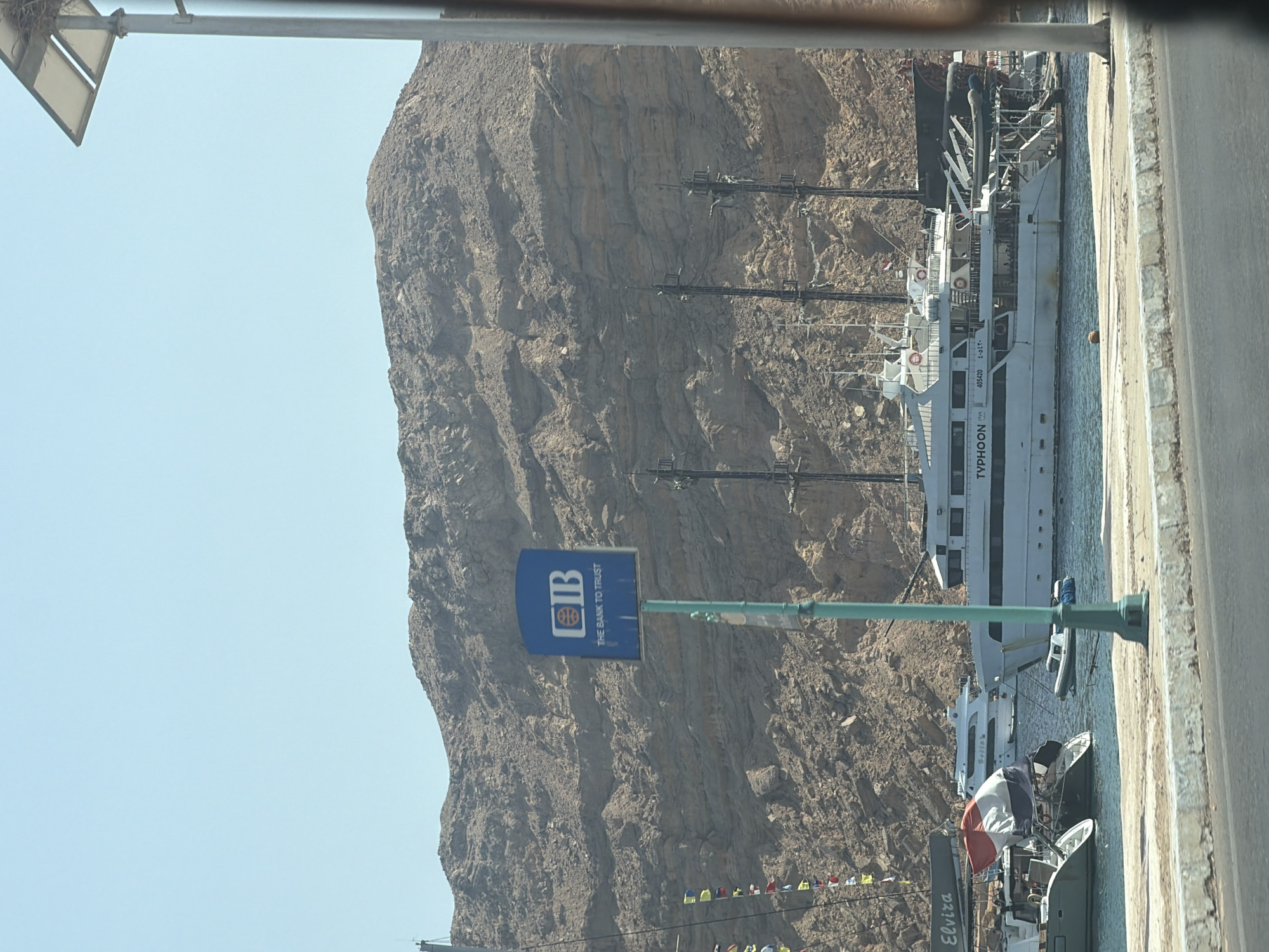 The image depicts a harbor scene in Sharm El Sheikh, Egypt, with a focus on a large white yacht named "TYPHOON" docked in the foreground. Behind the yacht, a steep, rocky mountain range rises under a clear blue sky. To the left of the yacht, smaller boats and a catamaran are visible, one with the name "ELVIRA" emblazoned on it. A red, white, and blue flag is prominently displayed on the catamaran. A blue sign with the "CIB THE BANK TO TRUST" logo is positioned to the left of the yacht. In the distance, another white yacht is anchored. The water is calm, reflecting the clear sky. The scene suggests a sunny day in a coastal tourist area.