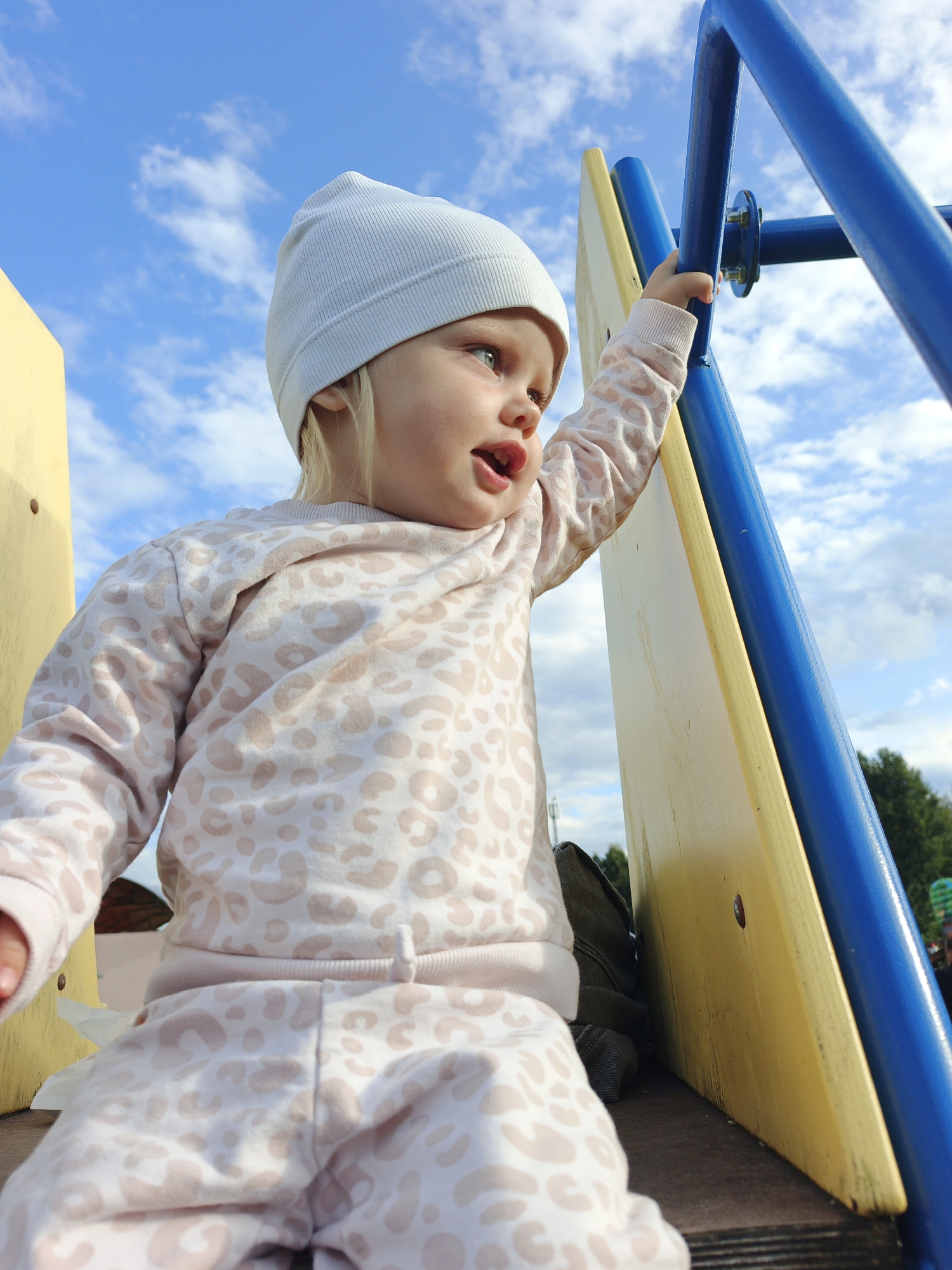 A low-angle outdoor shot captures a young child, likely a toddler, on a playground. The child, wearing a light-colored beanie and a matching loungewear set with a leopard-print pattern, is positioned on a wooden platform. The child's right hand grasps a blue metal bar, part of the playground equipment. The background consists of a bright blue sky with scattered white clouds, and the tops of green trees are visible in the distance. The playground structure features yellow and blue elements. The lighting suggests it is daytime.