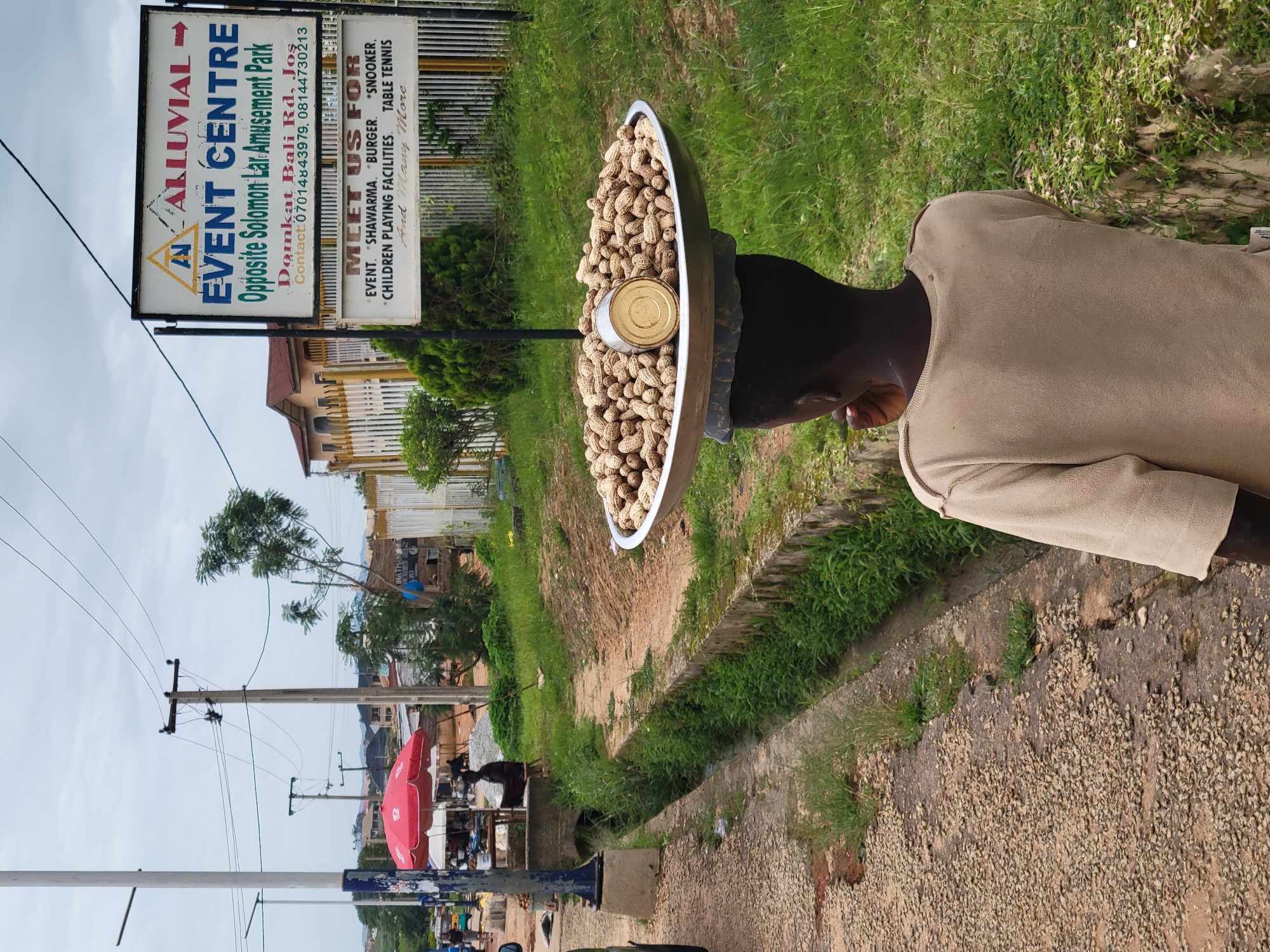 A young person is captured from behind, carrying a large, circular metal tray filled with peanuts balanced on their head. A tin can is placed in the center of the peanut mound. The person wears a light brown, long-sleeved shirt. They are positioned on a gravelly pathway next to a concrete drainage ditch overgrown with lush green grass and weeds. In the background, a white sign with black lettering is visible. The sign reads "ALLUVIAL EVENT CENTRE Opposite Solomon Lar Amusement Park" and lists "Domkat Bali Rd, Jos" as the contact address. Below this, another section of the sign states "MEET US FOR EVENT. *SHAWARMA. *BURGER. *SNOOKER. CHILDREN PLAYING FACILITIES. TABLE TENNIS. And Many More". Further in the background, there are buildings, a red umbrella, and utility poles with overhead wires, suggesting an urban or semi-urban setting within the city of Jos, Nigeria. The sky is overcast.