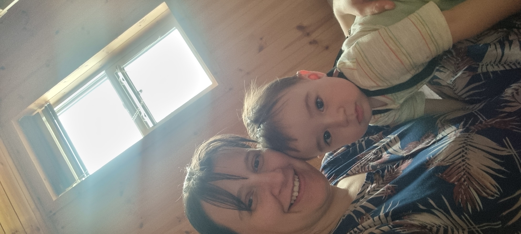 A selfie depicts a woman and a baby indoors. The woman, with short dark hair and a wide smile, is positioned at the bottom of the frame. Above her is a baby, lying on her chest, looking directly at the camera. The baby has dark hair and is wearing a light green shirt with stripes. The woman is wearing a blue shirt with a tropical leaf pattern. A window with closed blinds is visible in the upper left corner, suggesting daylight. The room has wooden walls. The location is in Dunnae-myeon, South Korea.