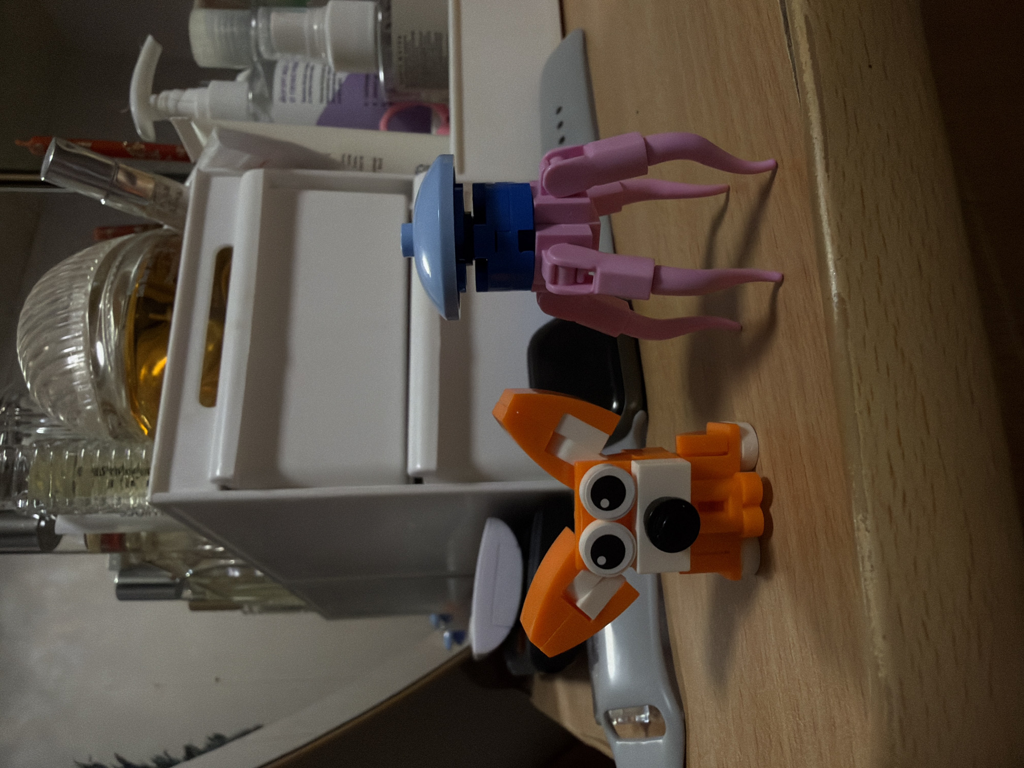 The image depicts a still life composition on a light wooden surface. Foreground items include an orange and white Lego fox and a blue and pink Lego jellyfish resting on a gray Apple Watch band. In the background, there is a partially visible white storage basket filled with various toiletries and perfume bottles. The environment suggests an indoor setting, possibly a bathroom shelf or vanity. The location is not discernible from the image and cannot be specified as Izhevsk, Russia, or any other city.