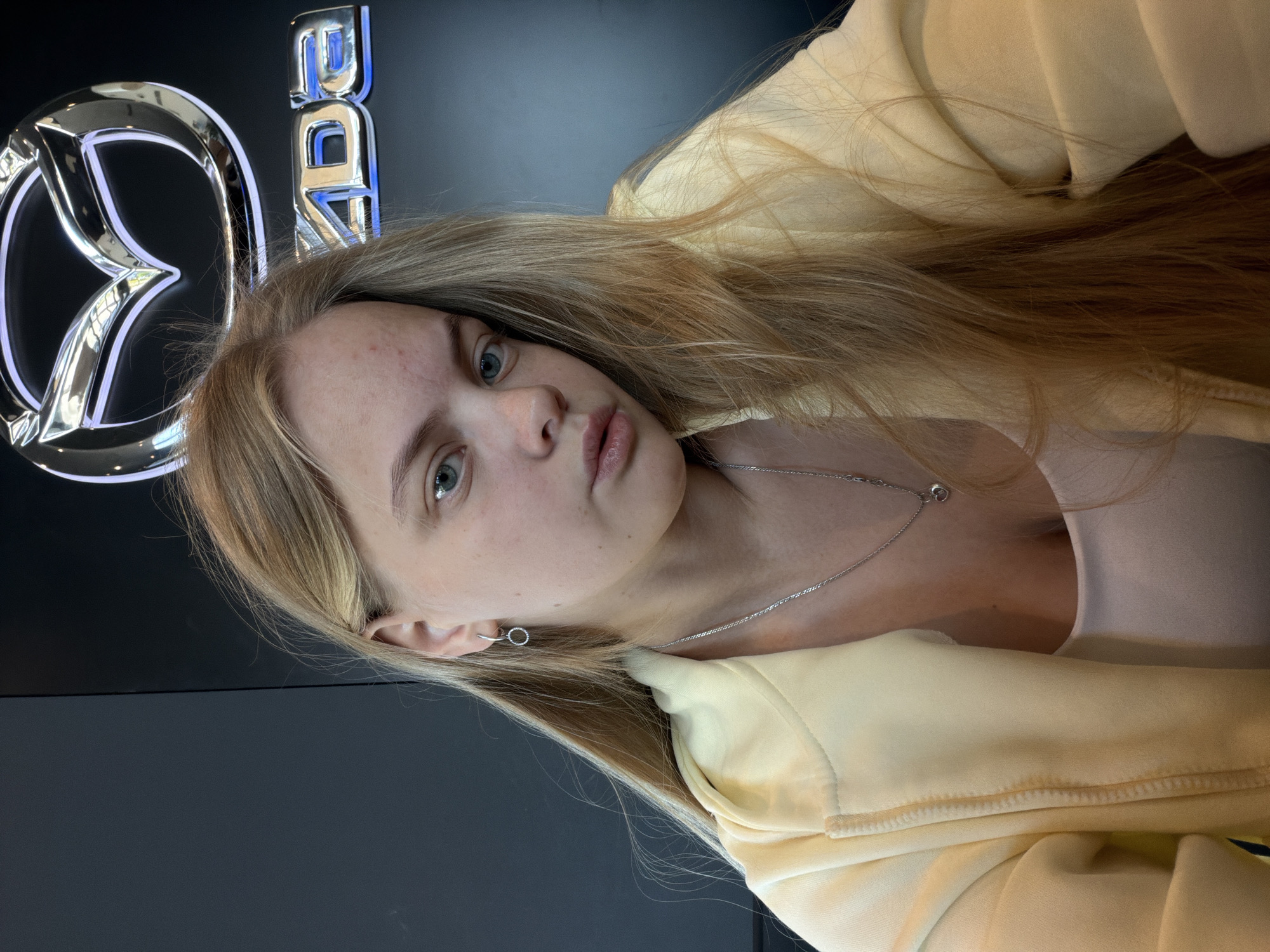 A young, light-skinned woman with long blonde hair is centered in the frame. She is wearing a light yellow hooded jacket and a thin silver necklace. She has light-colored eyes and appears to be looking directly at the camera with a slight pout. In the background, partially visible on the left, is a Mazda logo illuminated in blue and silver. The scene appears to be indoors, possibly in a car dealership. There is no further evidence to suggest this image was taken in the city of Izhevsk, Russia.