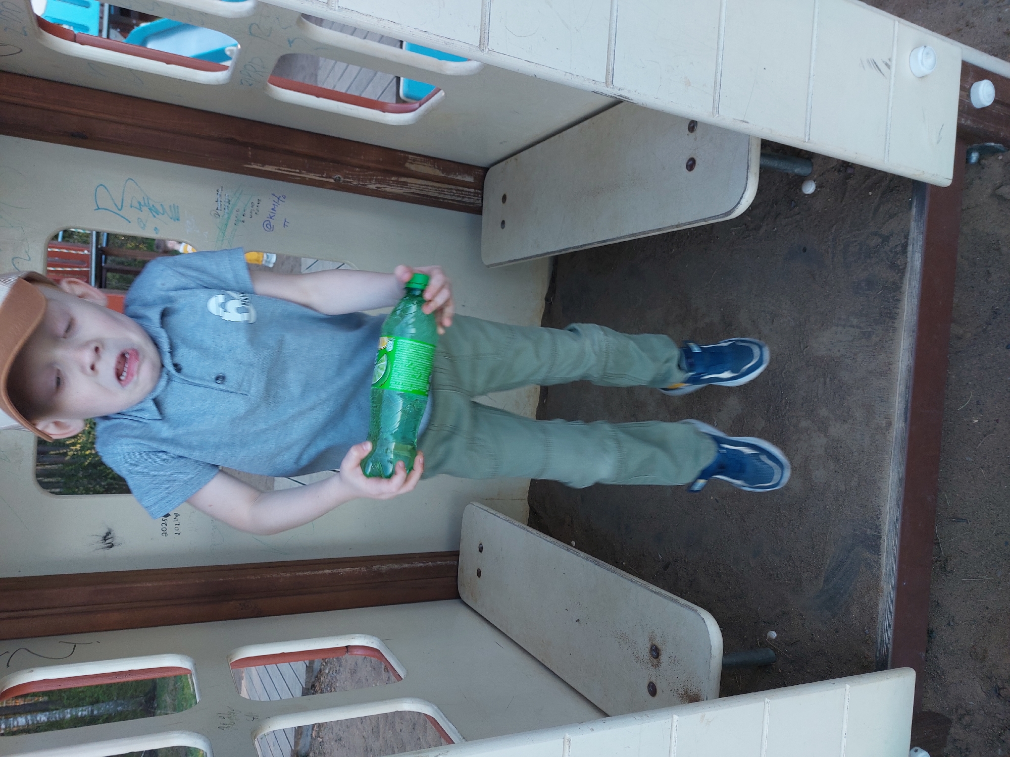 A young boy with a light complexion, wearing a brown baseball cap, light blue collared shirt, and green pants, is holding a green bottle of soda while standing inside a play structure. He appears to be in a park, presumably located in the city of Chaikovsky, Russia. The structure has a white and brown frame with open window-like spaces. The flooring and the surrounding ground is a mix of sand and dirt. A segment of the playground border can be seen on the right side of the frame. The image appears to be taken from an elevated, tilted perspective.
