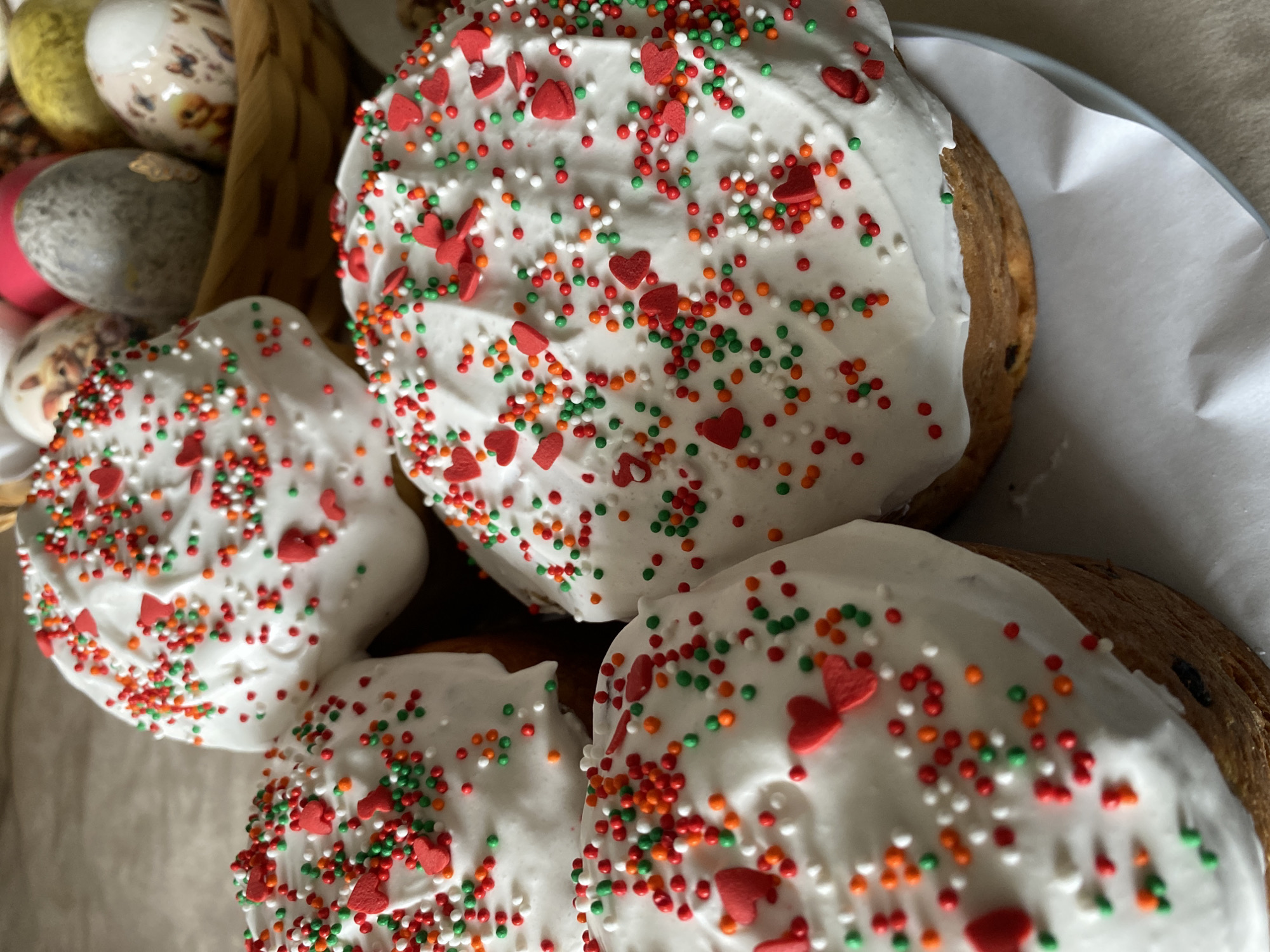 The image shows a close-up of several Easter cakes, also known as kulich, decorated with white icing and colorful sprinkles, including red hearts, green, white, orange, and red round sprinkles. Some decorative Easter eggs are visible in a basket in the upper left corner. The cakes are arranged closely together, with some on a plate lined with white paper. The image does not reveal the location or the city of Chaikovsky, Russia.