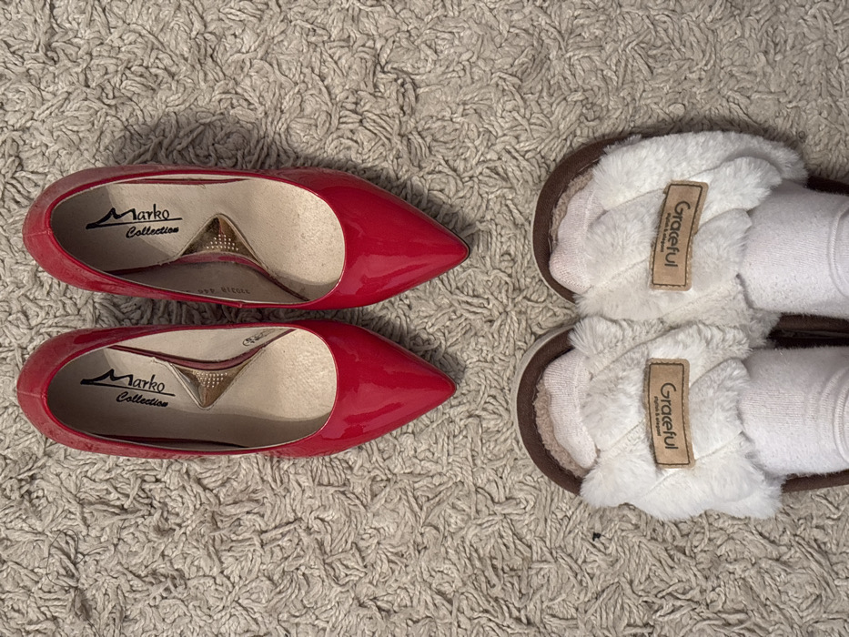 The scene, situated in the city of Velikiye Luki, Russia, depicts footwear arranged on a light-colored, textured carpet. In the upper half of the frame, a pair of red, pointed-toe, patent leather high-heeled pumps are visible. Their insoles display "Marko Collection" branding with a distinctive gold-colored insert. Below these pumps, a person's feet, wearing white socks, are observed inside a pair of brown and white furry slippers. Each slipper is adorned with a tan-colored tag featuring the word "Graceful".