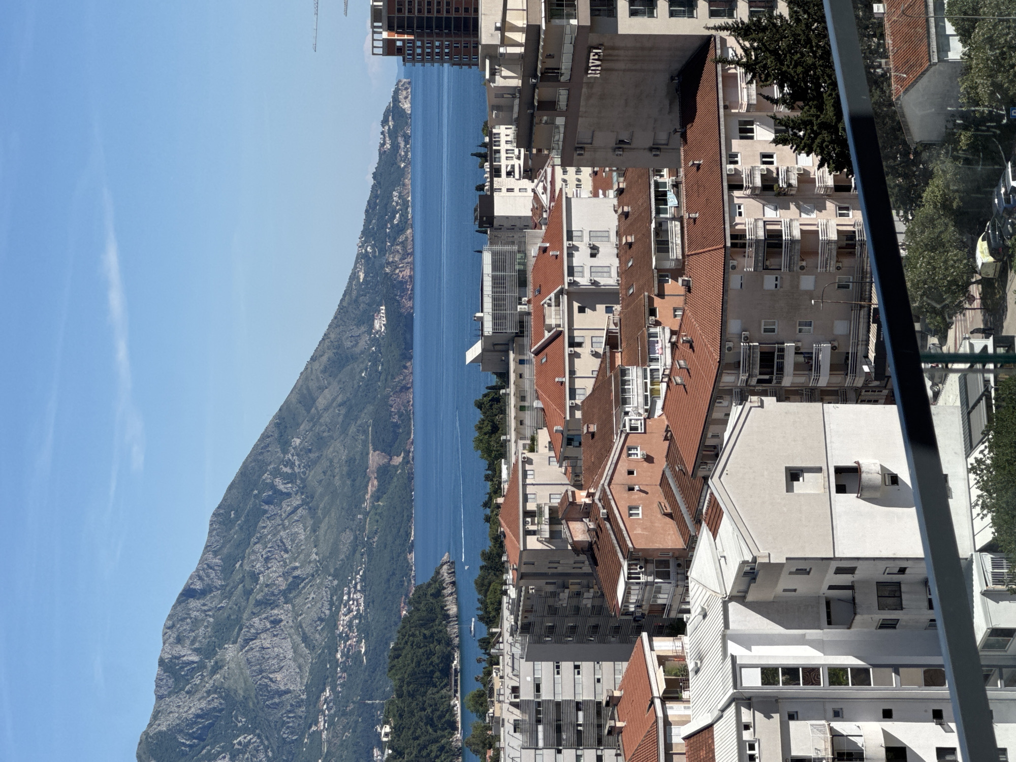 The image presents a high-angle, somewhat distorted, view of Budva, Montenegro. The left side of the frame shows a mountain range with a rugged terrain, transitioning into the blue sea. Several buildings and coastal vegetation are visible near the shore. The right side depicts an urban landscape of Budva, with closely packed buildings of varying heights and architectural styles. Most buildings are multi-story structures with red-tiled roofs. Air conditioning units are also visible on many buildings. A glimpse of a street with trees and parked cars can be seen on the far right. The overall view indicates a dense urban environment set against a backdrop of natural scenery, typical of coastal towns.