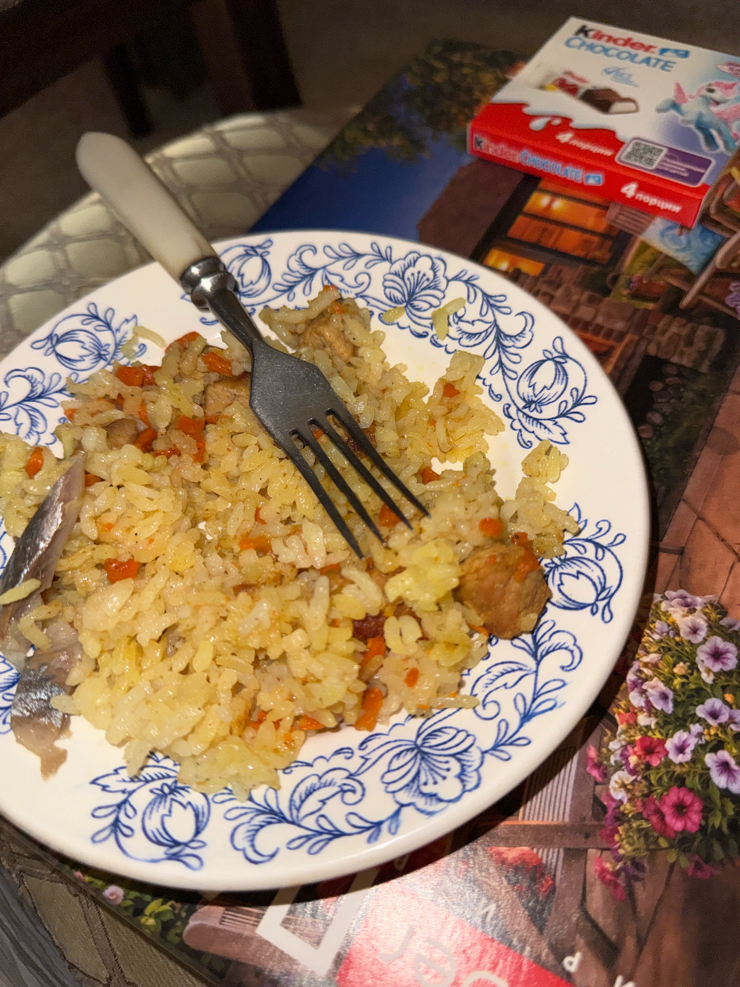 A close-up, high-angle, indoor shot shows a white plate with a blue floral pattern filled with rice, carrots, and meat. A fork with a white handle is partially submerged in the food. To the right of the plate, a red box of Kinder Chocolate is visible. The background appears to be a table covered with a patterned surface, possibly a magazine or placemat, featuring an image of potted flowers and a building. The setting is likely a home in Zavolzhye, Russia.