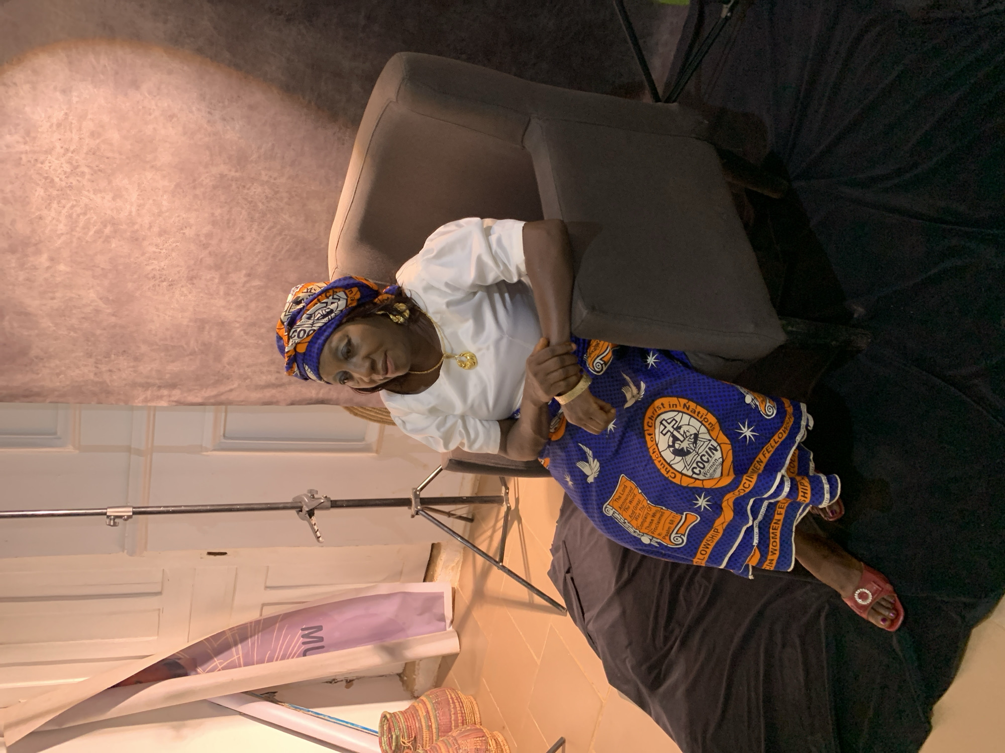 An overhead shot captures a middle-aged woman seated in a plush, dark grey armchair. She is adorned in traditional Nigerian attire: a white, puffed-sleeve blouse and a vibrant, blue skirt featuring an intricate orange and white pattern. The skirt is embellished with biblical verses, including "The Lord Announces The Word" and "Psalm 68:11". A matching headwrap, predominantly orange and blue, frames her face. She wears gold hoop earrings and a gold pendant necklace. Her hands are clasped in her lap, and she wears red open-toed sandals. A black cloth is draped over her legs and extends to the floor, partially obscuring a black beanbag chair. In the background, to the left, a white door stands open, revealing a roll of purple banner with white text, and two woven baskets. A tall, silver tripod stands to the right of the door. The wall behind the woman has a textured, pinkish-grey finish, with a circular light projection creating a spotlight effect. The scene appears to be a studio set, possibly in Jos, Nigeria, given the cultural attire and the hint of local elements.