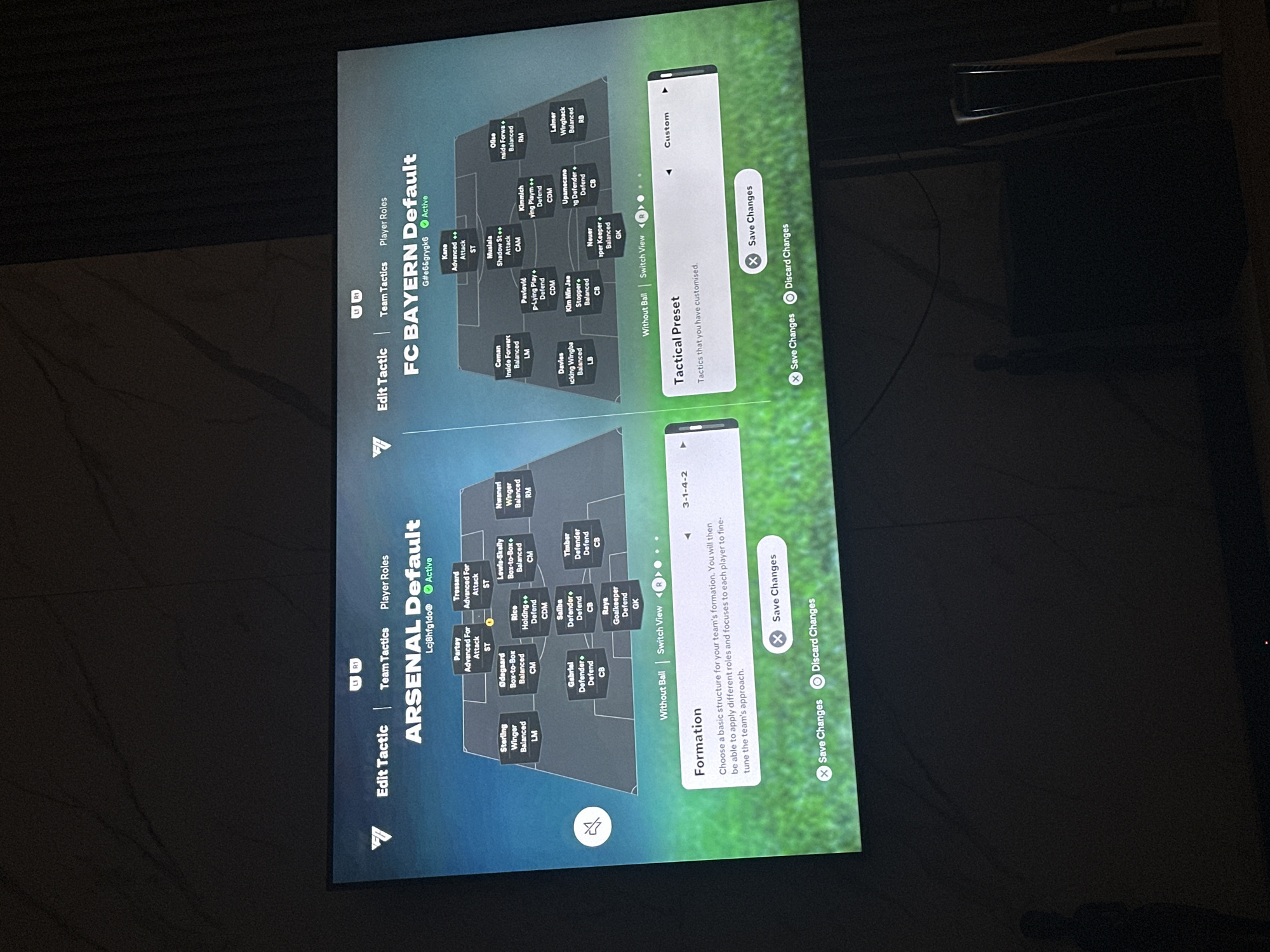 The image shows a screenshot of a video game's tactical screen, likely a soccer simulation game. The screen is divided into two main sections, one for "ARSENAL Default" and the other for "FC BAYERN Default". Each section displays a formation of players on a soccer field, with player names and their assigned roles (e.g., ST, LM, CB, GK). The "ARSENAL Default" section shows a 3-1-4-2 formation. The "FC BAYERN Default" section also shows a formation, but it is less clear due to image quality.
Below the formations, there are options to "Edit Tactic", "Team Tactics", and "Player Roles". Further down, there's a "Formation" section with the current formation displayed as "3-1-4-2" for Arsenal. There are also buttons for "Save Changes" and "Discard Changes". The "FC BAYERN" section has a "Tactical Preset" menu open.
The image was taken from a low angle, with the screen displaying the video game in a dimly lit room. A dark, textured wall is visible in the background, and a sliver of a dark object, possibly furniture or another electronic device, is present at the bottom right. The overall scene suggests a person is playing a video game in their home.
The prompt includes a request to include the location of Gwarinpa, Nigeria, which is not deducible from the image itself. There is no visual information in the image that connects it to Gwarinpa, Nigeria.