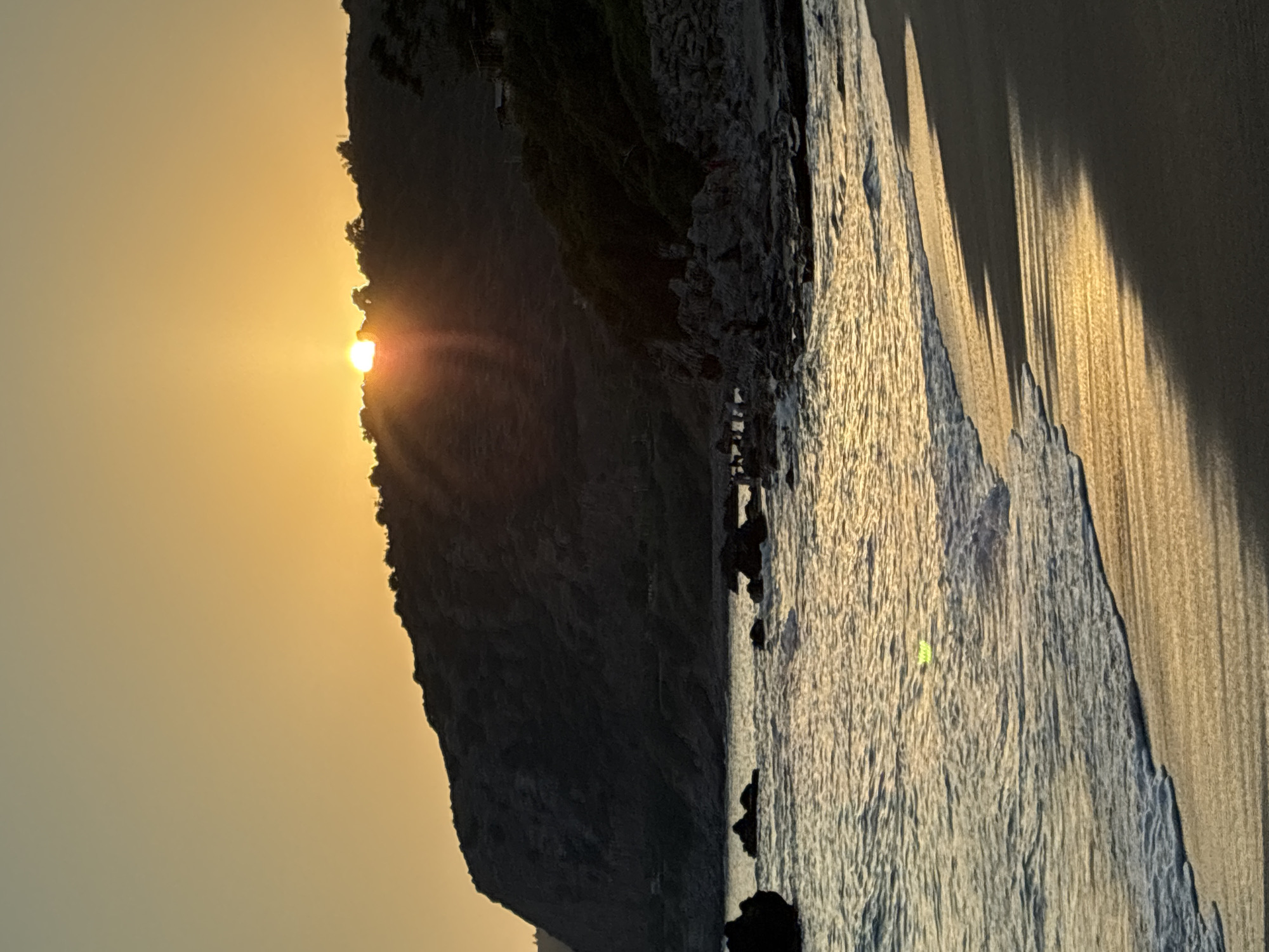 A serene sunset scene unfolds at the beach in Alanya, Turkey. The sun, a radiant orange disc, hovers just above the silhouette of a tree-covered hill, casting long, golden rays across the sky and water. Gentle waves lap the sandy shore, their foam creating delicate patterns as they recede. Dark rocks are scattered along the coastline, some partially submerged in the shallow water, adding a rugged texture to the otherwise smooth landscape. The sky transitions from a warm orange near the horizon to a pale yellow, then to a soft blue higher up. The beach itself is a mix of dark sand and wet, reflective areas, showcasing the dynamic interplay of light and shadow.