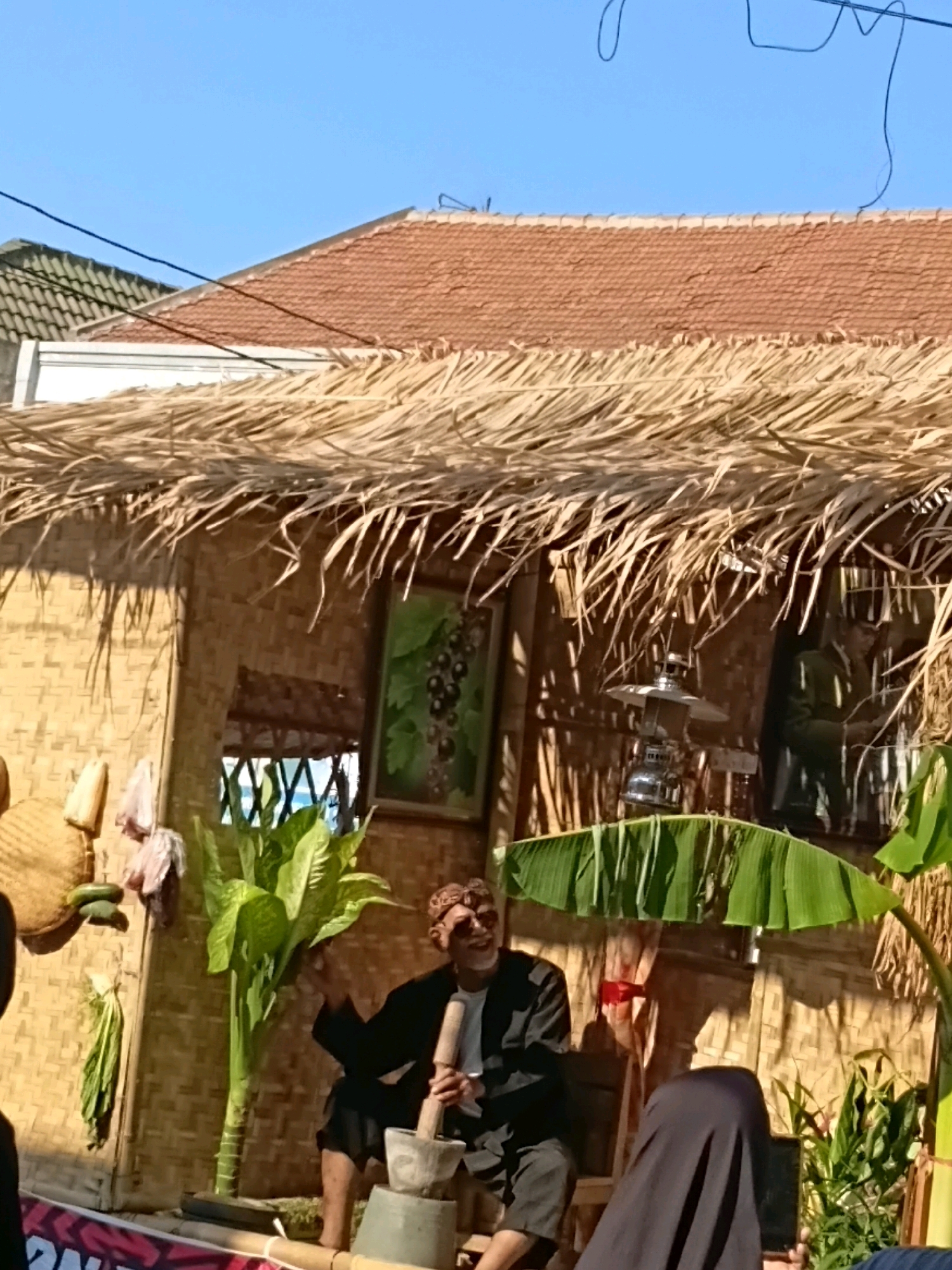 In Wadungasri, Indonesia, a man dressed in traditional attire, including a patterned head covering and a black jacket, sits in front of a wall made of woven bamboo. He is actively engaged in crushing something in a mortar and pestle, with a large green plant to his left. The scene is set under a thatched roof, with a framed picture of dark berries hanging on the bamboo wall. Other decorative elements such as dried corn, other plants, and a metal lantern are visible. To the right, a person in black clothing partially obstructs the view. The background reveals rooftops and a clear blue sky, suggesting an outdoor setting on a sunny day.