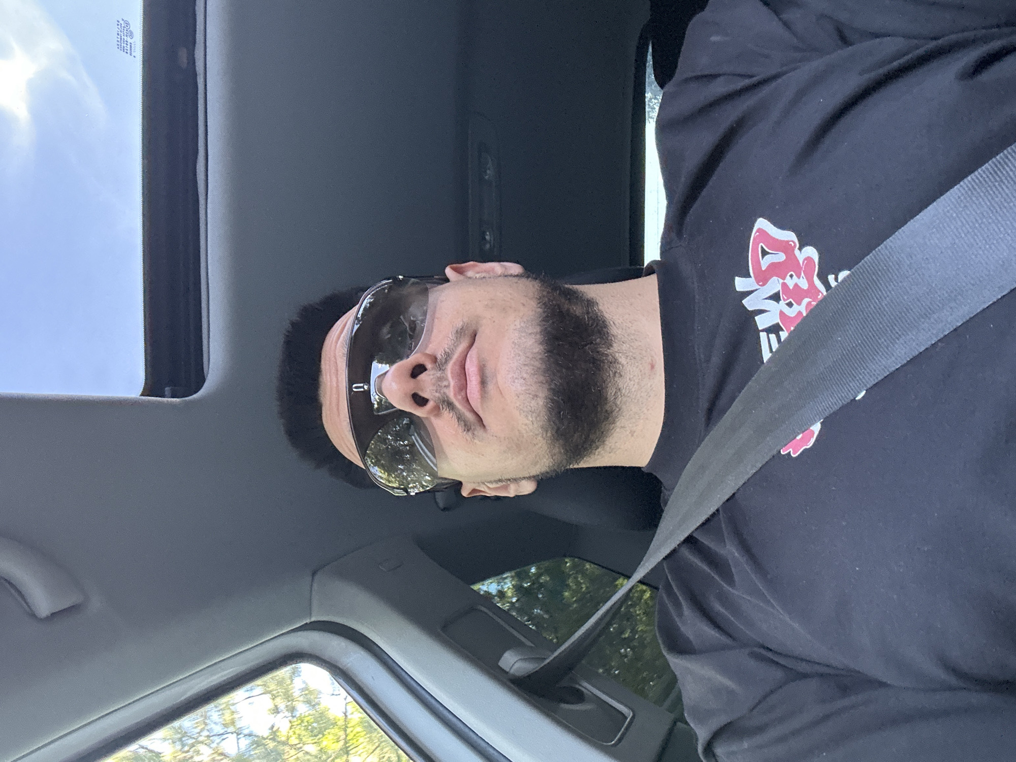 The media file shows a person wearing sunglasses and a black t-shirt with a red and white graphic design, possibly a logo or artwork. The person is inside a car, as evidenced by the seatbelt, car interior details like the sunroof, and the view of trees and foliage through the windows. The image is taken from a low angle, looking up at the person. The lighting suggests it is daytime, with sunlight coming through the sunroof. The person's expression, with closed eyes and slightly parted lips, could indicate relaxation or a moment of repose. The background visible through the windows suggests a rural or suburban setting with green trees. The overall mood is casual and relaxed.