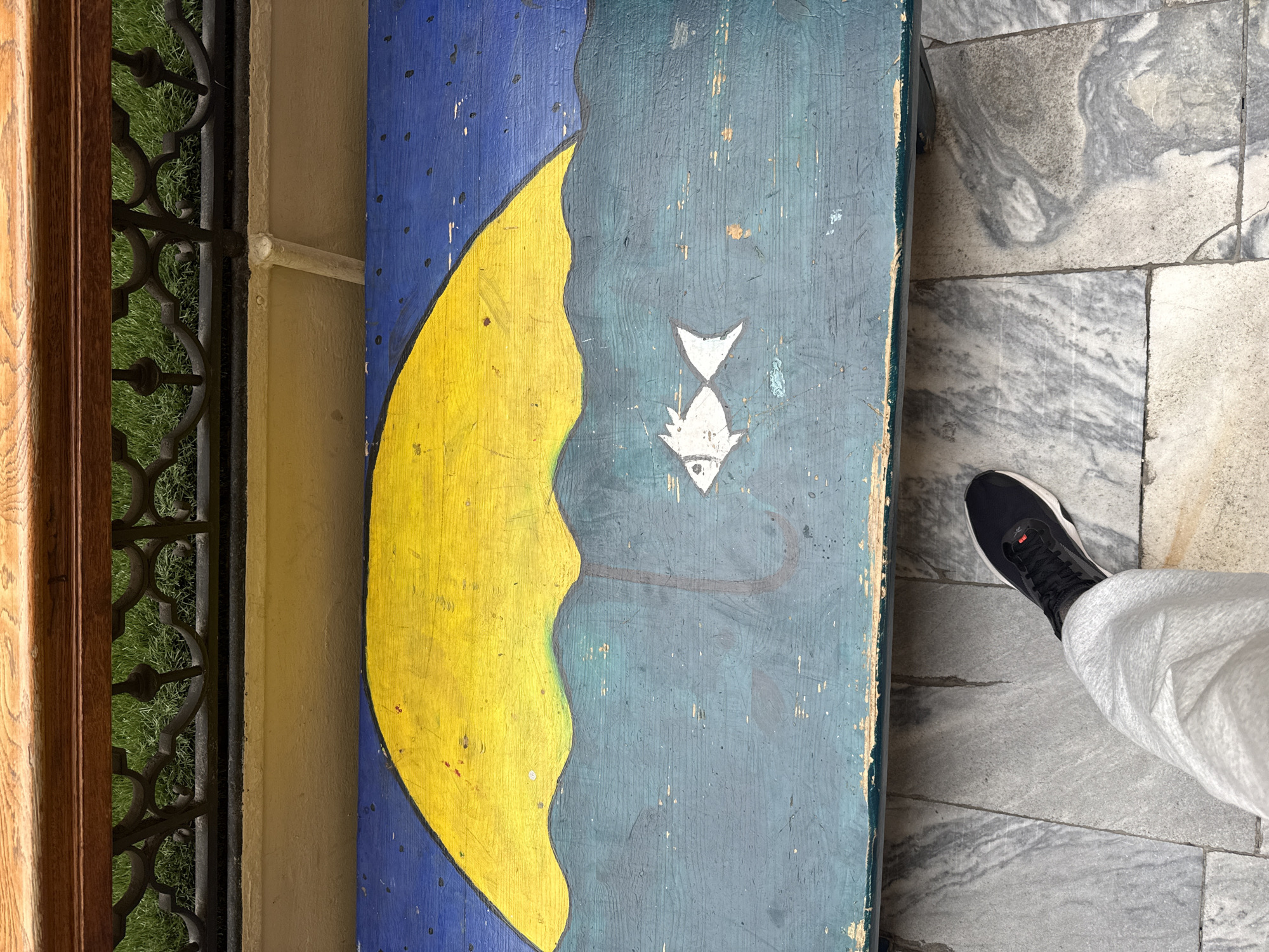 The image shows a painted wooden panel with a crescent moon and a fish, standing on a marble-tiled floor. A person's foot in a black and white shoe is partially visible on the right. A decorative iron gate is visible to the left of the panel, with a view of green grass behind it. The painting has a naive style with visible brushstrokes and signs of wear and tear. The setting is possibly a building interior. The location in the city of Moscow, Russia is not clear from the image.