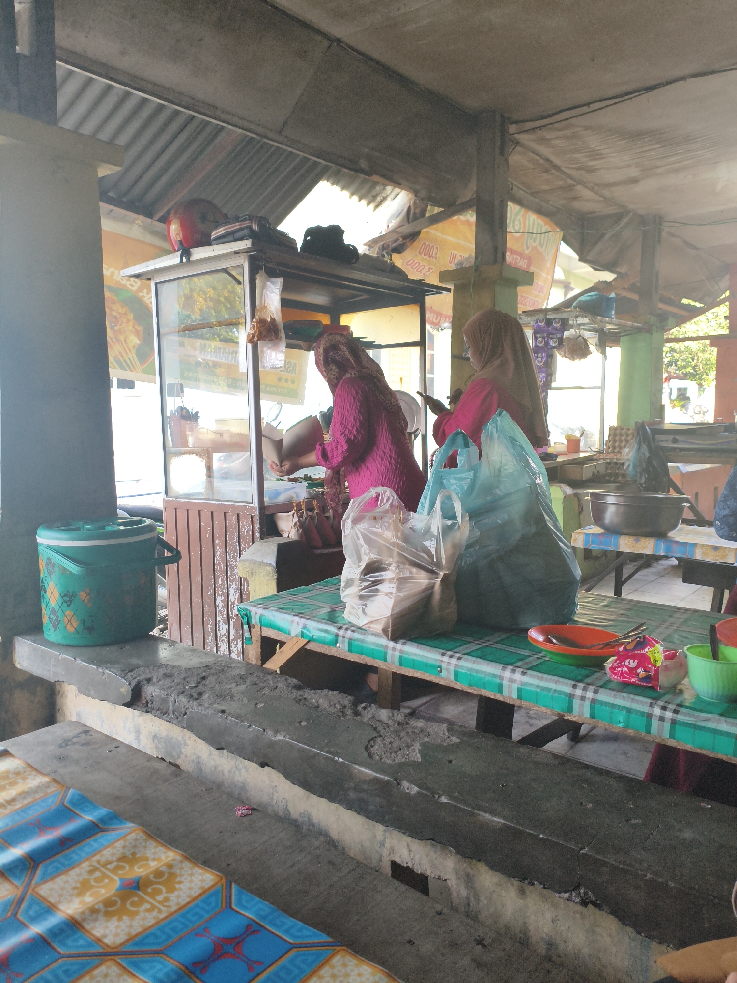 The image depicts an outdoor food stall in the city of Tebing Tinggi, Indonesia. Two women, both wearing hijabs, are working at the stall. One woman, in a pink headscarf and pink shirt, is serving food from a glass display case. The other woman, in a brown hijab and a pink top, is standing and holding a smartphone. Various food items are visible in the display case and packaged in plastic bags on the table. A large green cooler is placed on a concrete ledge to the left of the stall. The stall is covered by a corrugated metal roof supported by wooden posts. The environment suggests a bustling marketplace or a roadside food vendor area.
