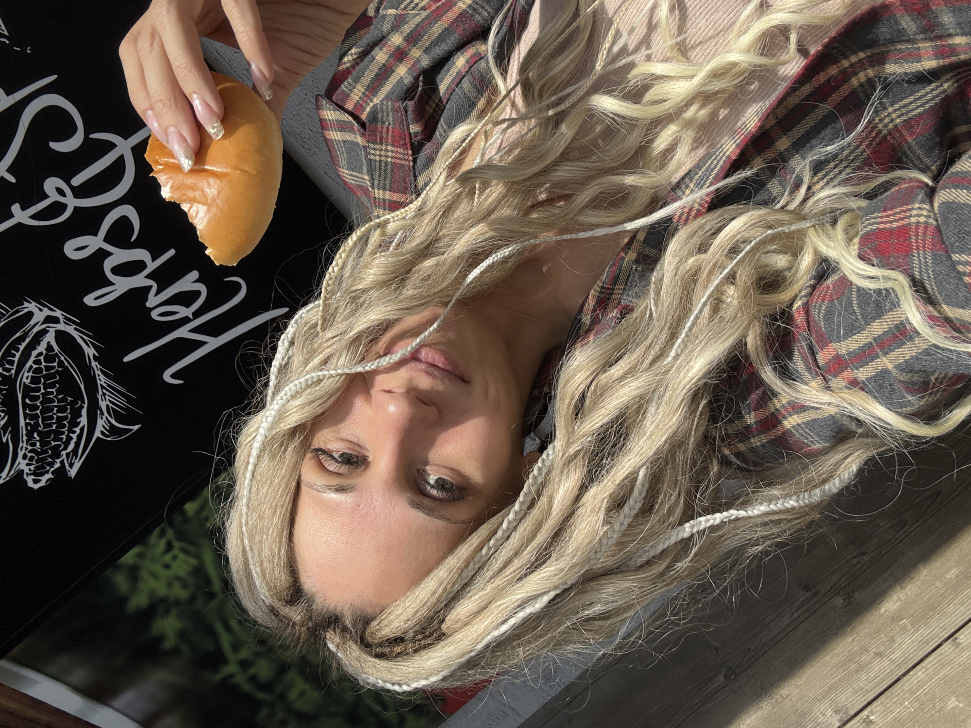 A woman with blonde hair with white braids interspersed is looking at the camera. She's wearing a tan shirt and a plaid flannel shirt. In her hand, she's holding a golden-brown bread roll, partially eaten. The location appears to be outdoors. There's a black sign with white lettering and an illustration of corn. Some green foliage is visible in the background. The presence of wooden planks indicates a possible deck or table. The location is not identifiable as Rusanovo, Russia from the visual evidence.