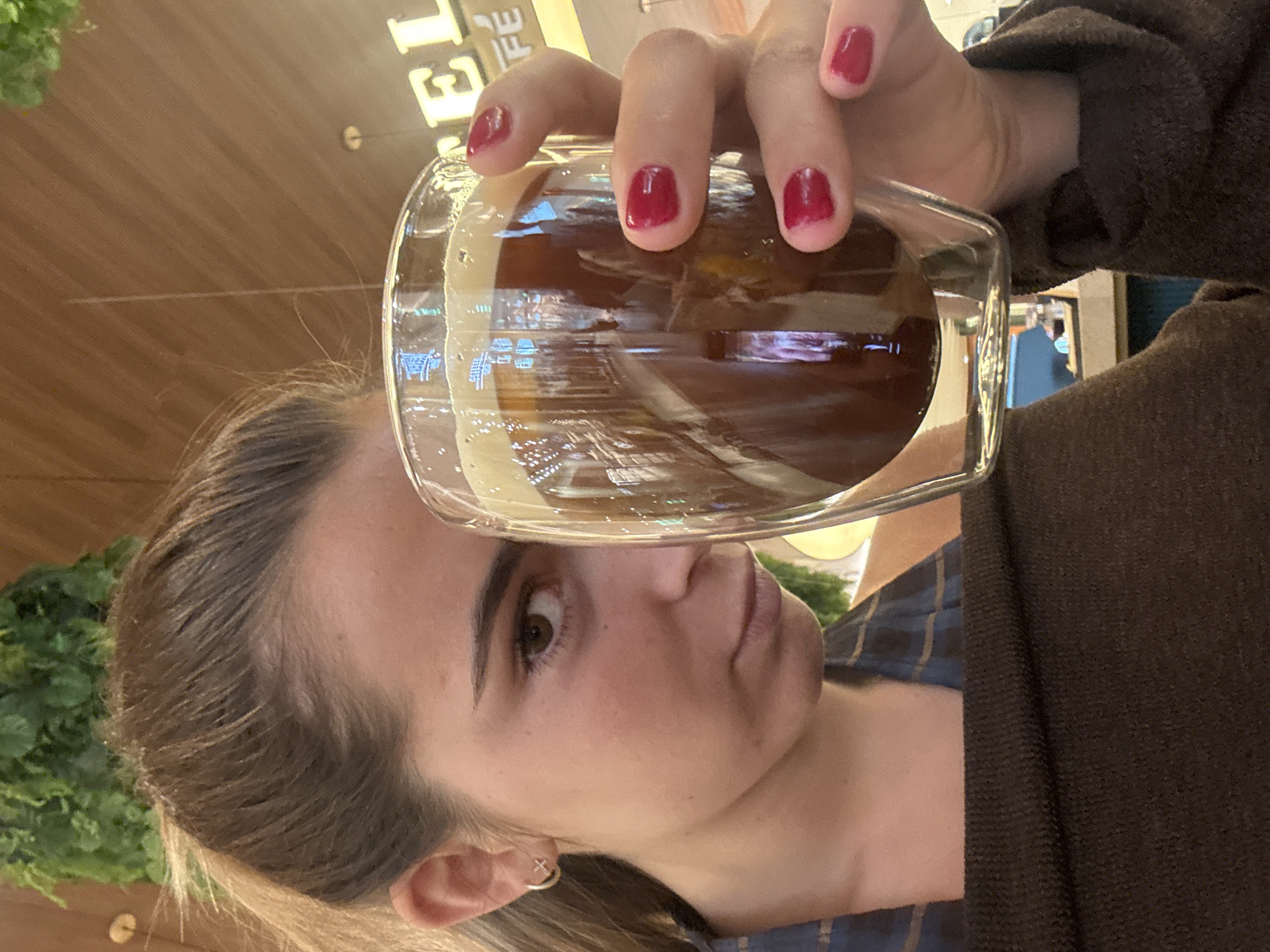 A woman with fair skin and brown hair is holding a glass of dark liquid, likely iced coffee, in front of her face. Her nails are painted a dark red color. She is wearing a plaid shirt and a brown sweater. She is looking at the camera with a neutral expression. In the background, there are green plants and a sign that says "CAFE." The scene is likely inside a café. The café may be located in Senhora da Hora, Portugal, but this is not evident from the image.