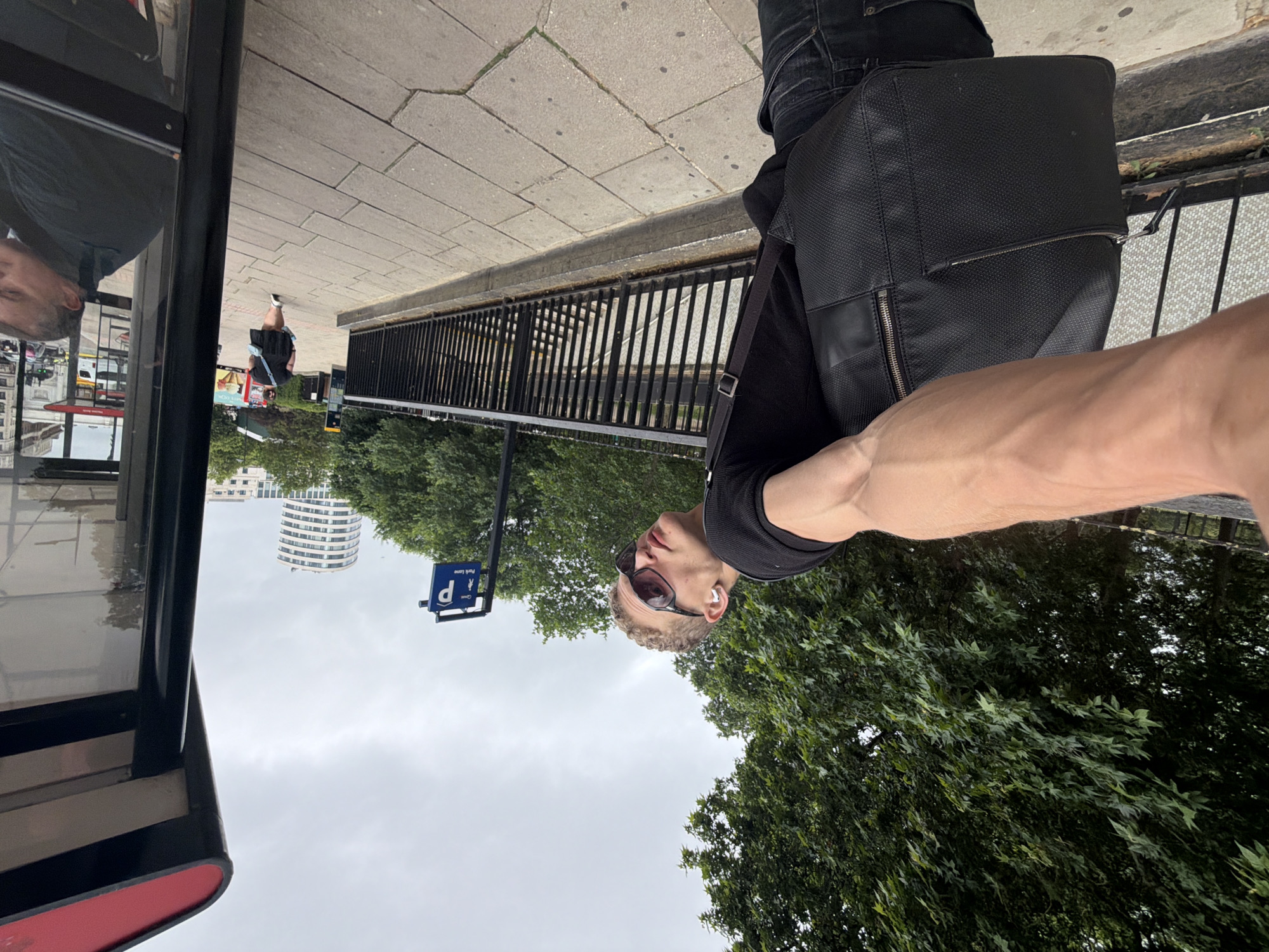 This is a low-angle, outdoor, eye-level shot taken from the perspective of a person standing at a bus stop in London, United Kingdom. The sky is overcast, and lush green trees are visible in the background, partially obscuring a cylindrical building. To the left, the red and black structure of the bus stop shelter is prominent. A blue parking sign with a white 'P' symbol and pictograms of people is mounted on a pole. The foreground features the arm and shoulder of the photographer, who is wearing a black t-shirt, sunglasses, and earbuds, and carrying a black messenger bag. A black metal railing runs along the right side of the frame, separating the bus stop area from a paved walkway with a subtle grid pattern. Further down the walkway, a person in a black dress and white sneakers is visible, walking away from the camera. In the reflection of the bus stop shelter glass, another person is seen reclining.