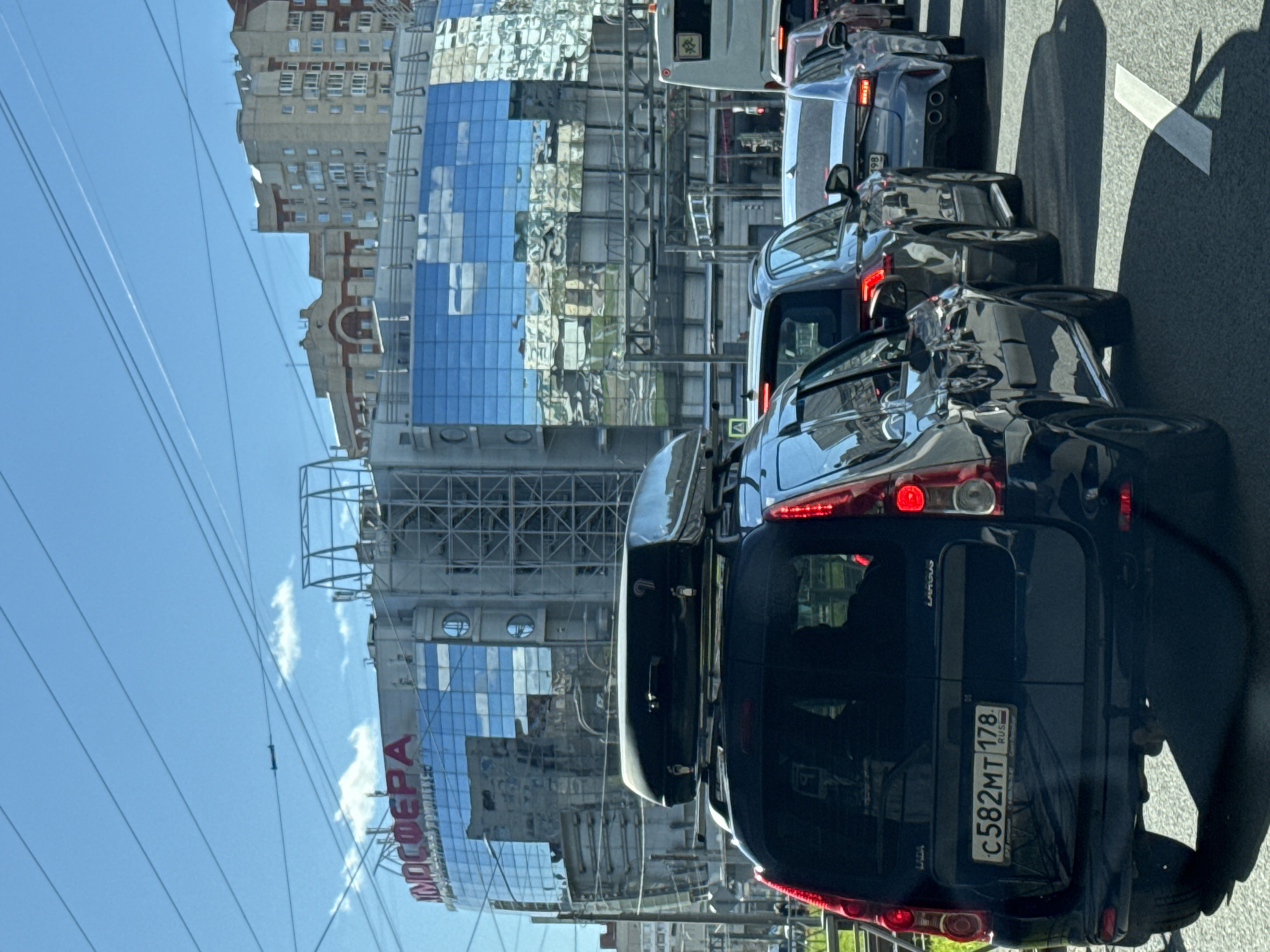 The image depicts a congested urban street scene in Saint Petersburg, Russia, with several vehicles in a line of traffic. A dark-colored Lada Largus van with a luggage carrier on top is prominent in the foreground, its license plate reading "C582MT 178," indicating it's registered in Saint Petersburg. Other cars are visible ahead. On the left side, a modern glass-faced building with the large word "МОСФЕРА" vertically stacked can be seen. Overhead power lines are visible. The clear blue sky suggests the scene is set on a sunny day.