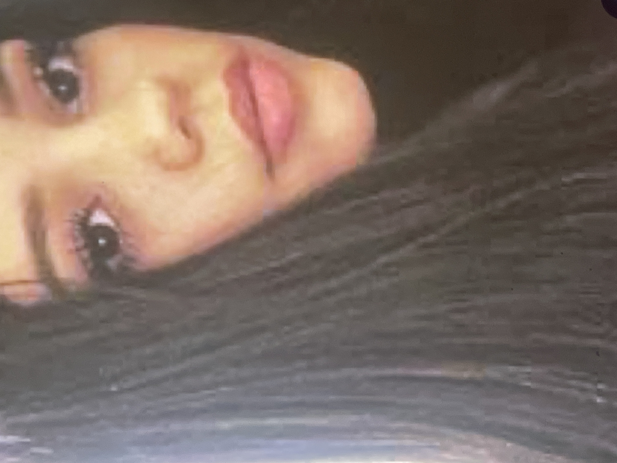 An intimate portrait features a young woman captured from the chest up, lying on her side with her head angled towards the viewer. Her long, dark brown or black hair is prominently displayed, fanned out behind her. Her facial features are visible, including both eyes which are open and adorned with dark lashes, a well-defined nose, and soft, parted lips that bear a natural pinkish-red color. Her skin tone is light to medium. The background is dark and indistinct, suggesting an indoor setting with soft, ambient lighting. The image exhibits a slightly grainy texture, enhancing its candid and natural feel. No specific activity is discernible beyond posing or resting. There is no visible text in the media file. The image is set in Poltava, Ukraine.