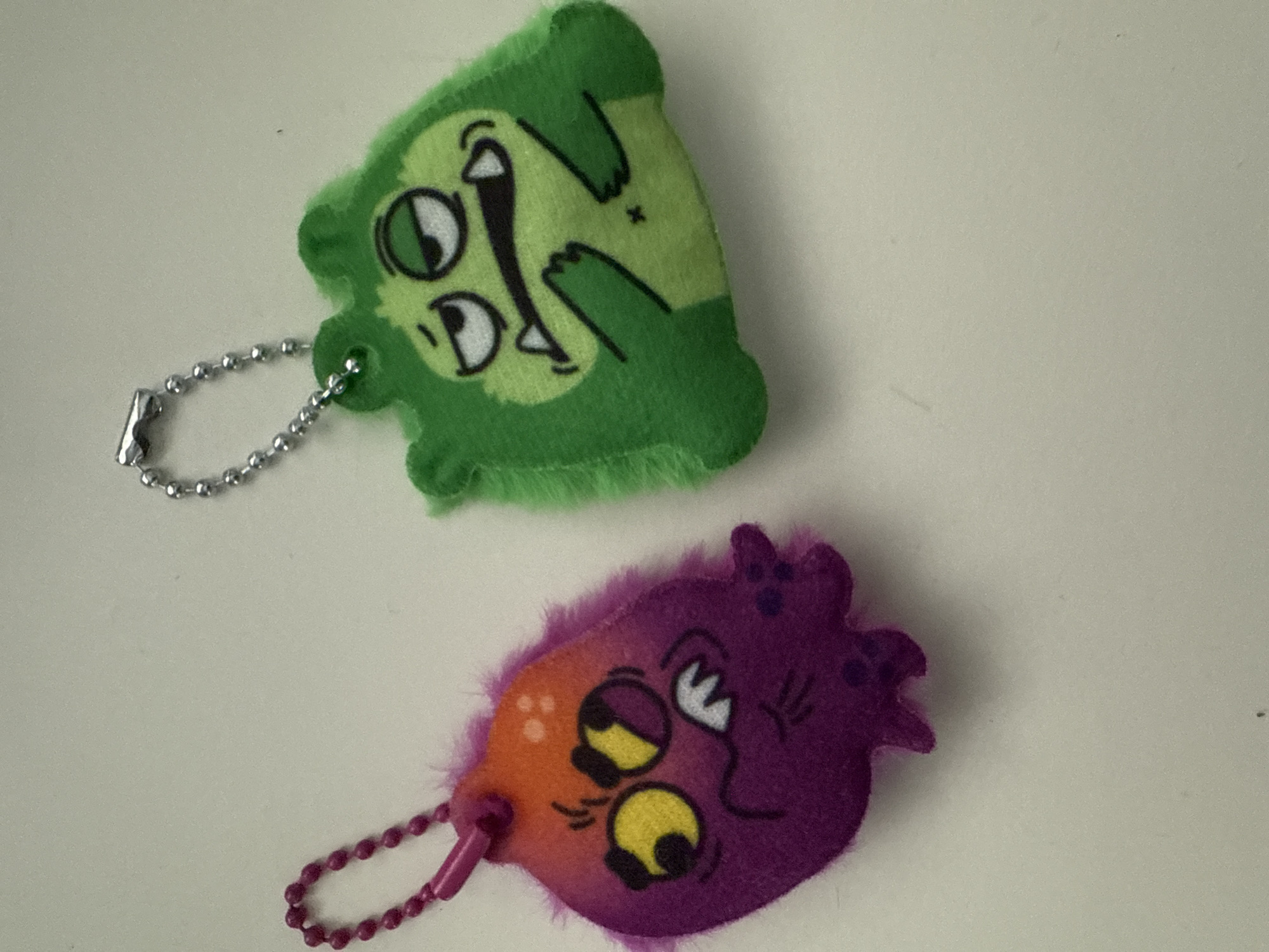 The image shows two cartoon-like plush keychains on a plain, likely white or light-colored surface. The top keychain is green and depicts a character with two eyes, an open mouth, and stylized limbs. It has a silver-colored ball chain attached. The bottom keychain is predominantly purple with some orange near the attachment point. It also features a cartoon character with two eyes and a similar whimsical design. It is attached to a purple ball chain. The image does not depict the location in the city of Votkinsk, Russia.