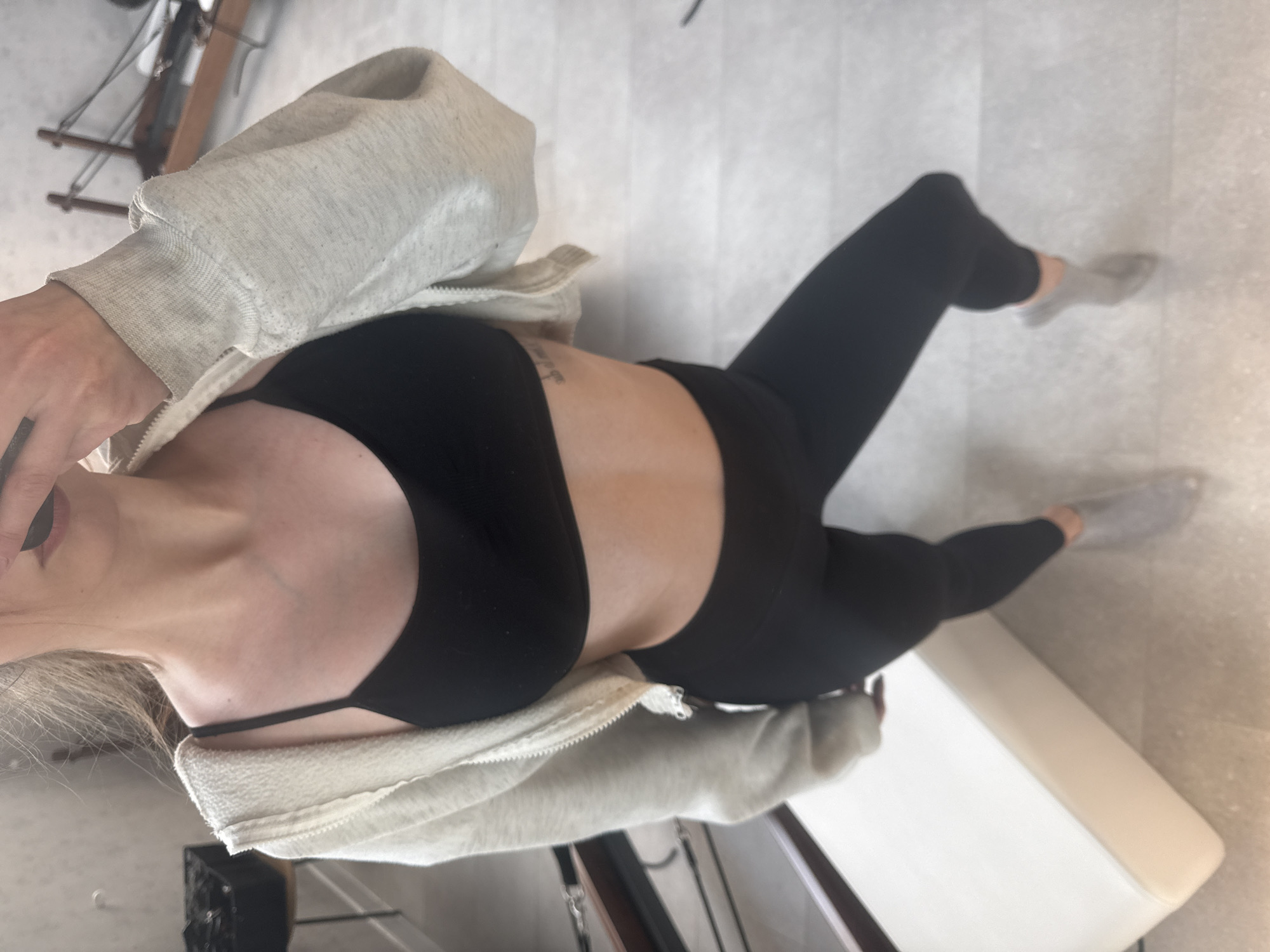 A woman is photographed from an overhead perspective. She is wearing a black sports bra, black leggings, and gray socks, and a light gray zip-up hoodie. A partial view of her face is visible, revealing makeup. A tattoo is noticeable on her torso. She appears to be standing indoors on a light-colored tile floor. Adjacent to her, there is a white upholstered bench and fragments of furniture can be seen in the background. The location cannot be determined to be Sofia, Bulgaria based on the visual information provided.