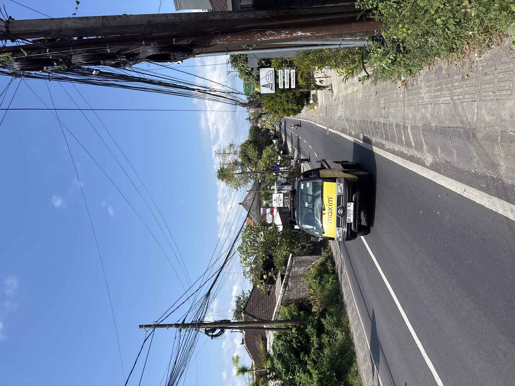 The image presents a vibrant daytime street scene in Kuta Selatan, Indonesia, captured under a clear blue sky with sparse white clouds. A paved road with a solid white line runs through the center, flanked by a textured concrete sidewalk on the right and lush tropical vegetation on both sides. Overhead, a dense tangle of black electrical and communication wires spans between sturdy utility poles.
A yellow and black Suzuki vehicle, featuring the "maxim" logo prominently on its front, is visible on the right side of the road, either parked or in motion. Its Indonesian license plate, "DK 8295 HJ," is clearly readable. Maxim is an app-based ride-hailing and delivery service, suggesting the vehicle's function. Further down the road, other vehicles, including a motorcycle, can be observed.
Along the right side of the street, amidst the abundant greenery, several signs are displayed. One sign features an illustration of a traditional temple structure and another lists various services: "Cooking class & Balinese dancing," "Coffee smoothies," and "Lunch, Dinner." These details, combined with the "DK" license plate code (associated with Bali), firmly place the scene within the Indonesian cultural context. Traditional buildings with dark tiled roofs are partially visible through the trees on the left side of the road. The bright sunlight and vibrant foliage suggest a warm, tropical climate.
