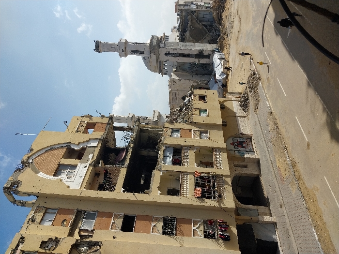 The image depicts a scene of extensive urban destruction in Khan Yunis, Palestinian Territory, under a partly cloudy sky. In the foreground and left mid-ground, a multi-story, light-tan residential building stands heavily damaged, with significant portions of its facade missing, exposing interiors. Numerous small impact marks are visible across its exterior. Balconies with white balustrades show hanging laundry. The ground floor features arched openings, one displaying graffiti.
To the right of this building, a large area is reduced to rubble and debris. A prominent mosque with an intact, tall, slender minaret and a dome is visible in the background, surrounded by damaged structures and piles of debris. White tarpaulin structures, possibly makeshift shelters, are situated among the rubble.
An asphalt road with white lane markings spans the lower and middle sections of the frame, bordered by a paved sidewalk on the left and dirt/rubble on both sides. Several individuals are present: one person in a yellow shirt walks in the center of the road, two dark-clad figures walk towards the lower right, and two to three smaller figures are visible further back near the tarpaulin structures. No direct interactions between individuals are discernible.