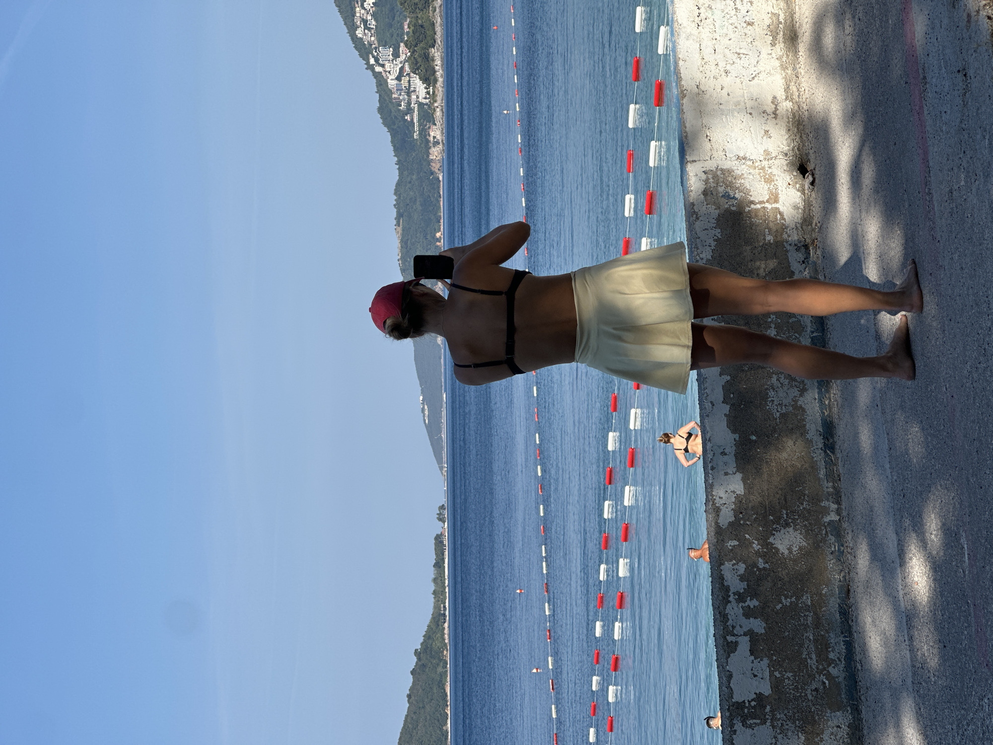 In Boreti, Montenegro, a young woman wearing a red baseball cap, a black bikini top, and a light yellow pleated skirt stands facing away from the camera, holding a smartphone in her right hand, seemingly taking a photograph of the Adriatic Sea. The sea is a deep blue, with a series of red and white buoys marking a swimming area. In the background, across the water, a hilly coastline is visible, dotted with white buildings. The foreground consists of a textured concrete wall and a paved surface with dappled sunlight. Several other people are in the water, some swimming and one appearing to be snorkeling. The sky is clear and blue.